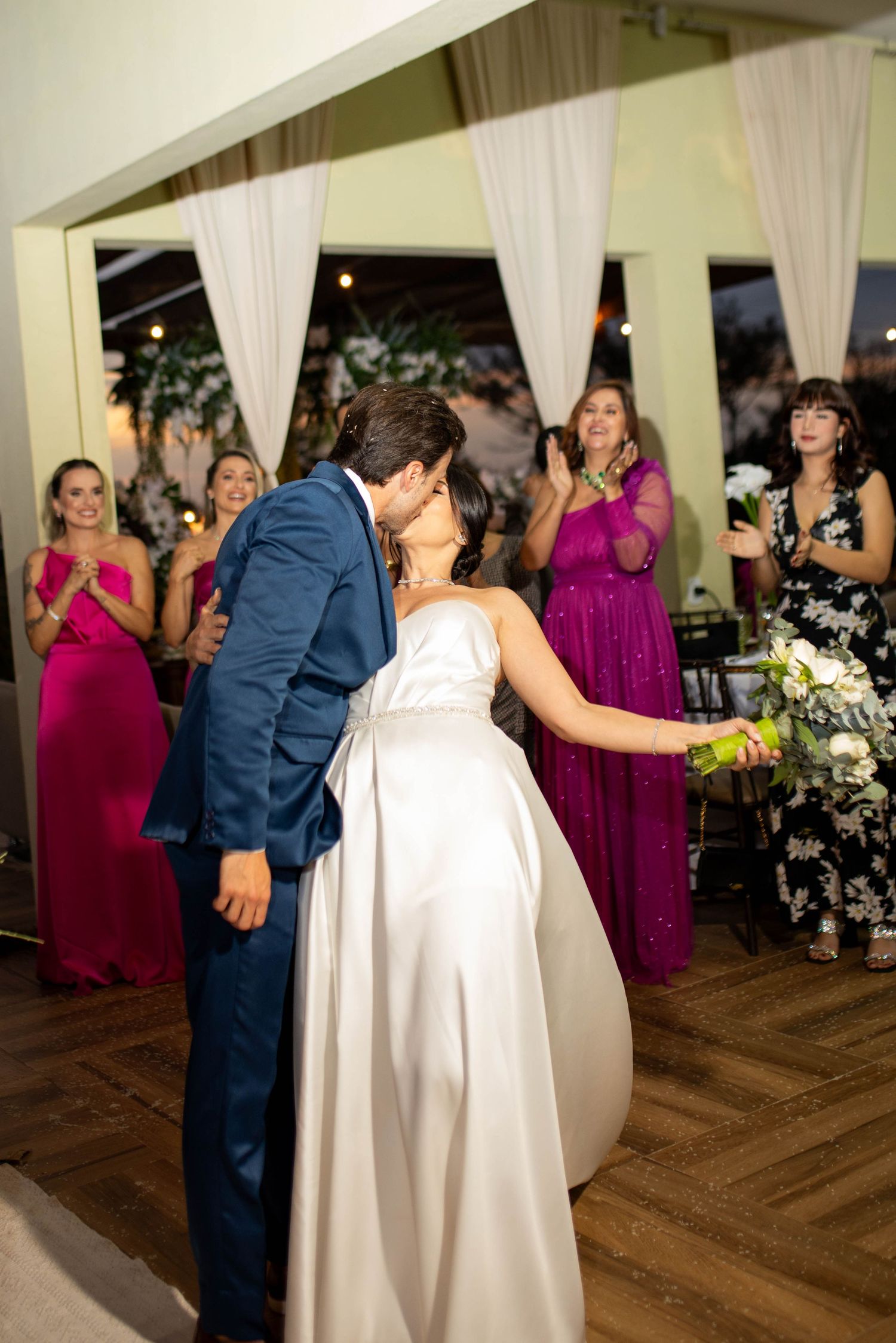 Bride and groom share a kiss on the dance floor while bridesmaids in pink and floral dresses applaud at wedding reception.