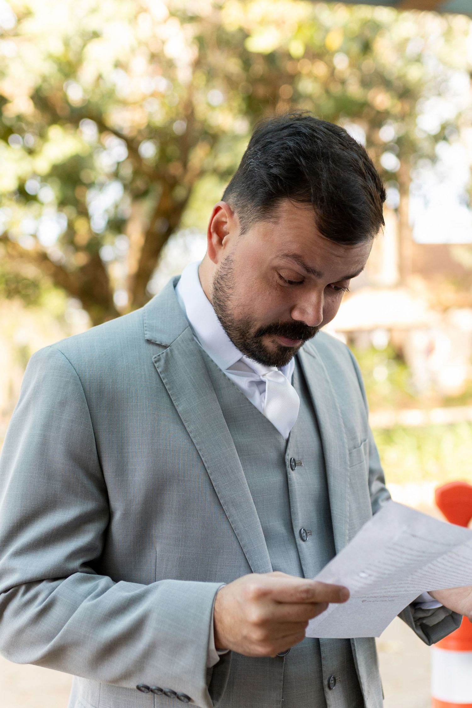Bearded man in gray suit reading document outdoors with blurred green trees in background.