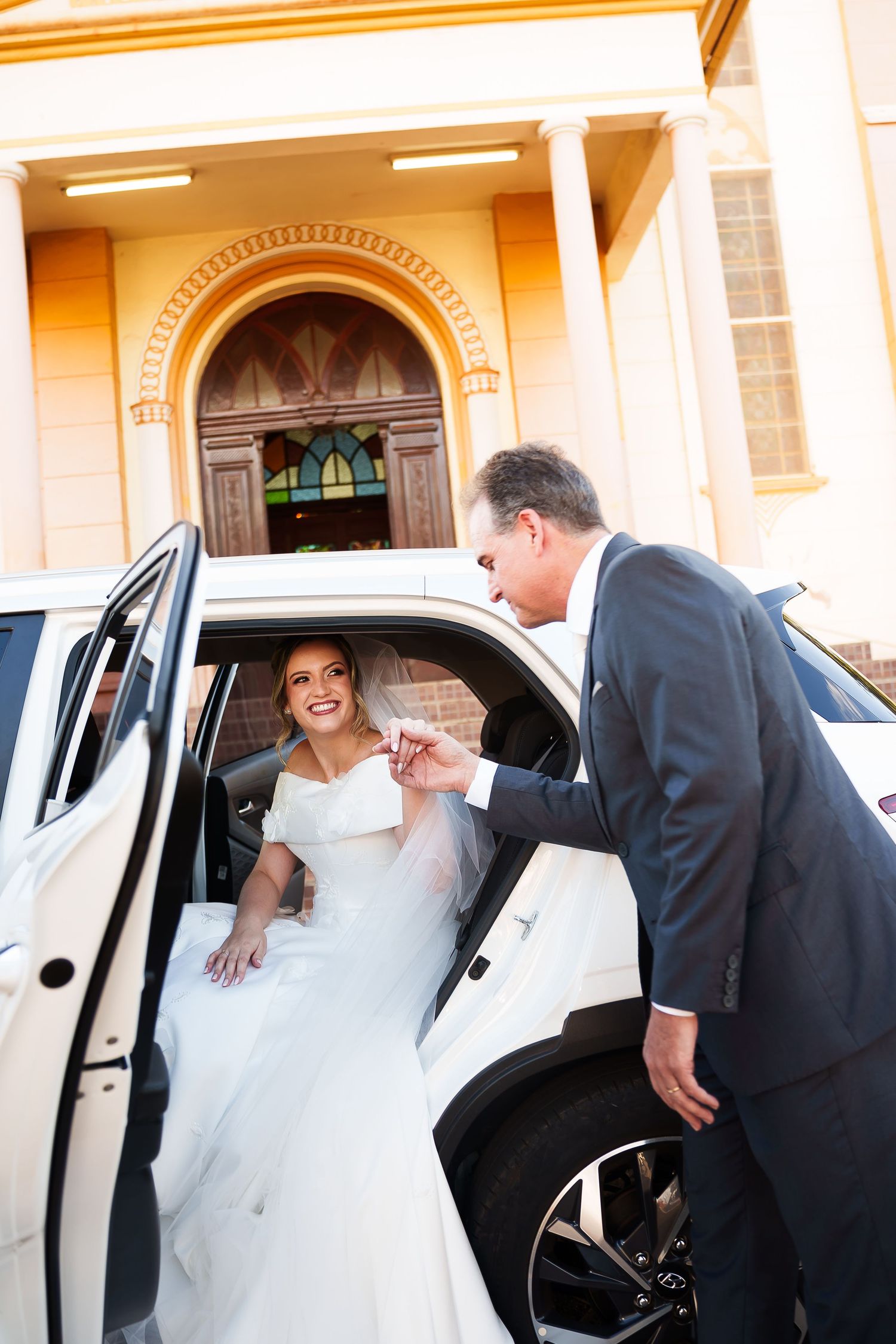 Groom helps bride exit white limousine outside church on wedding day.