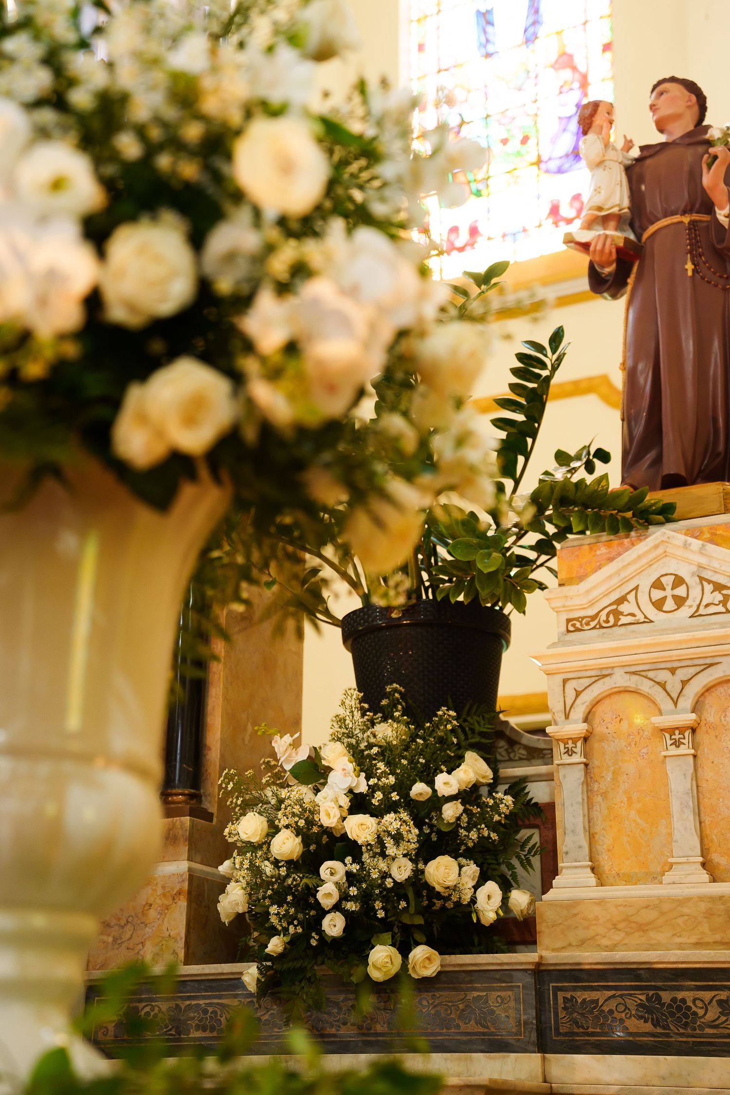Church altar adorned with white roses and a Saint Francis statue, bathed in warm light from a stained glass window.