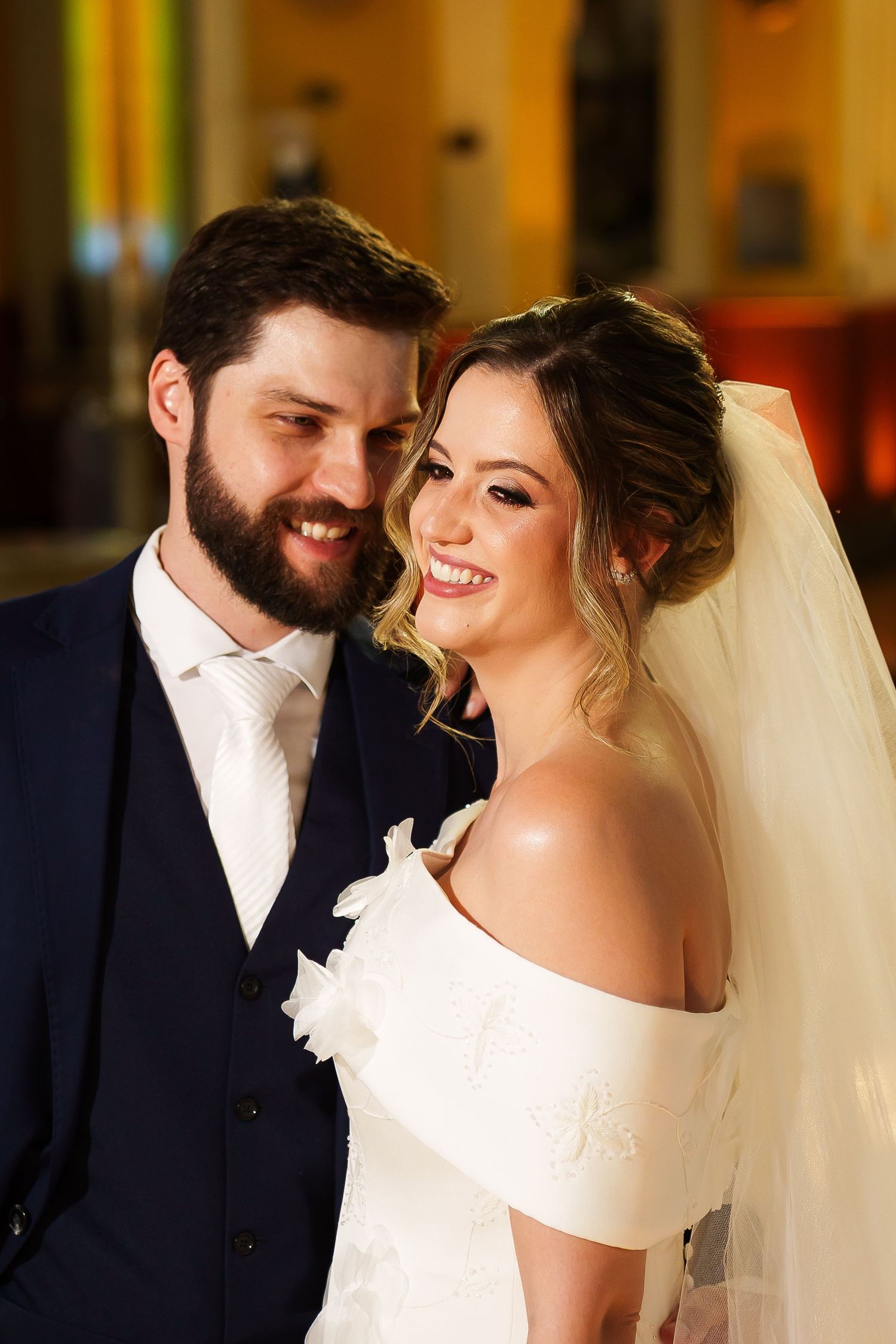 Smiling bride in white off-shoulder gown with veil and groom in navy suit posing together at wedding ceremony.