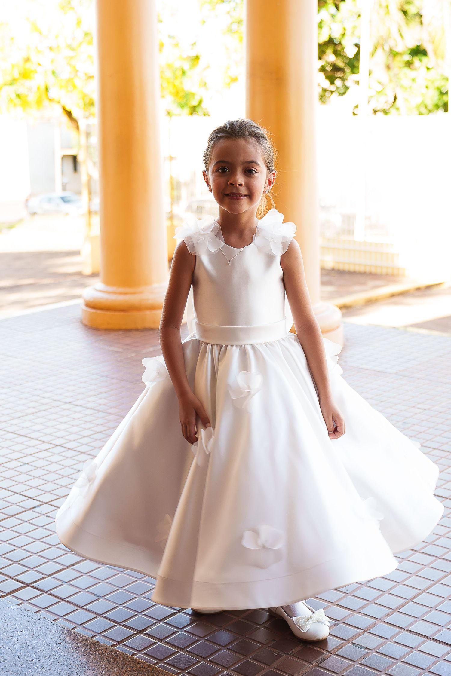 Young girl in white flower girl dress posing between yellow columns at outdoor ceremony venue.