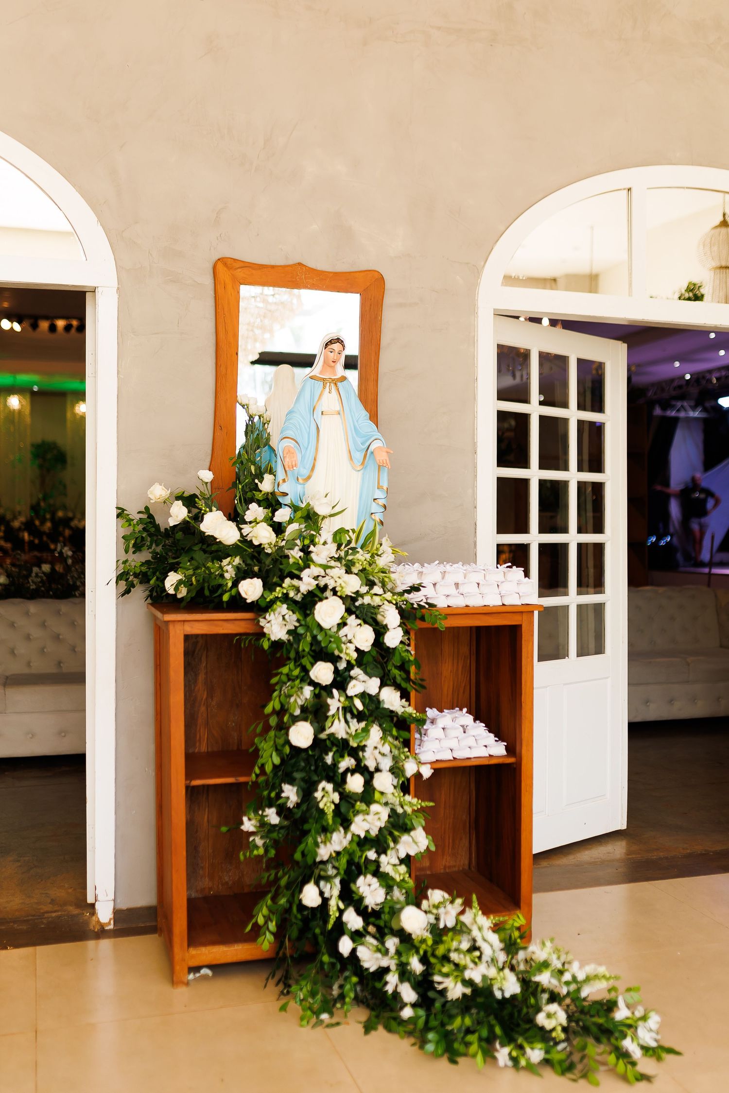 Wooden shelf with Virgin Mary statue, white floral arrangement, and small favors near arched doorways.