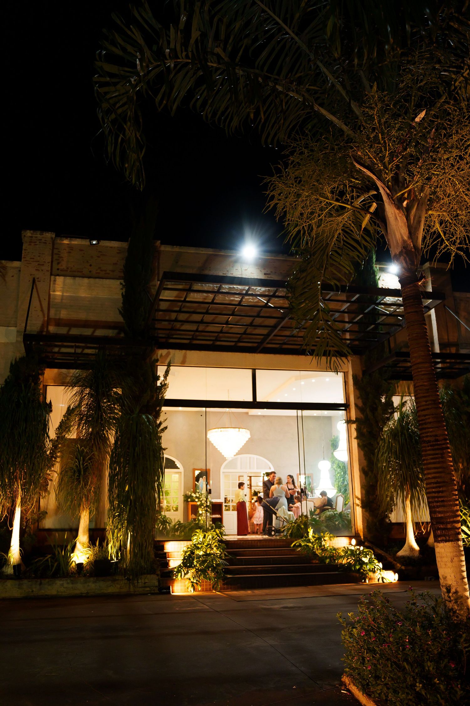 Elegant venue entrance lit at night with palm trees, glowing lights, and guests visible inside through large glass windows.