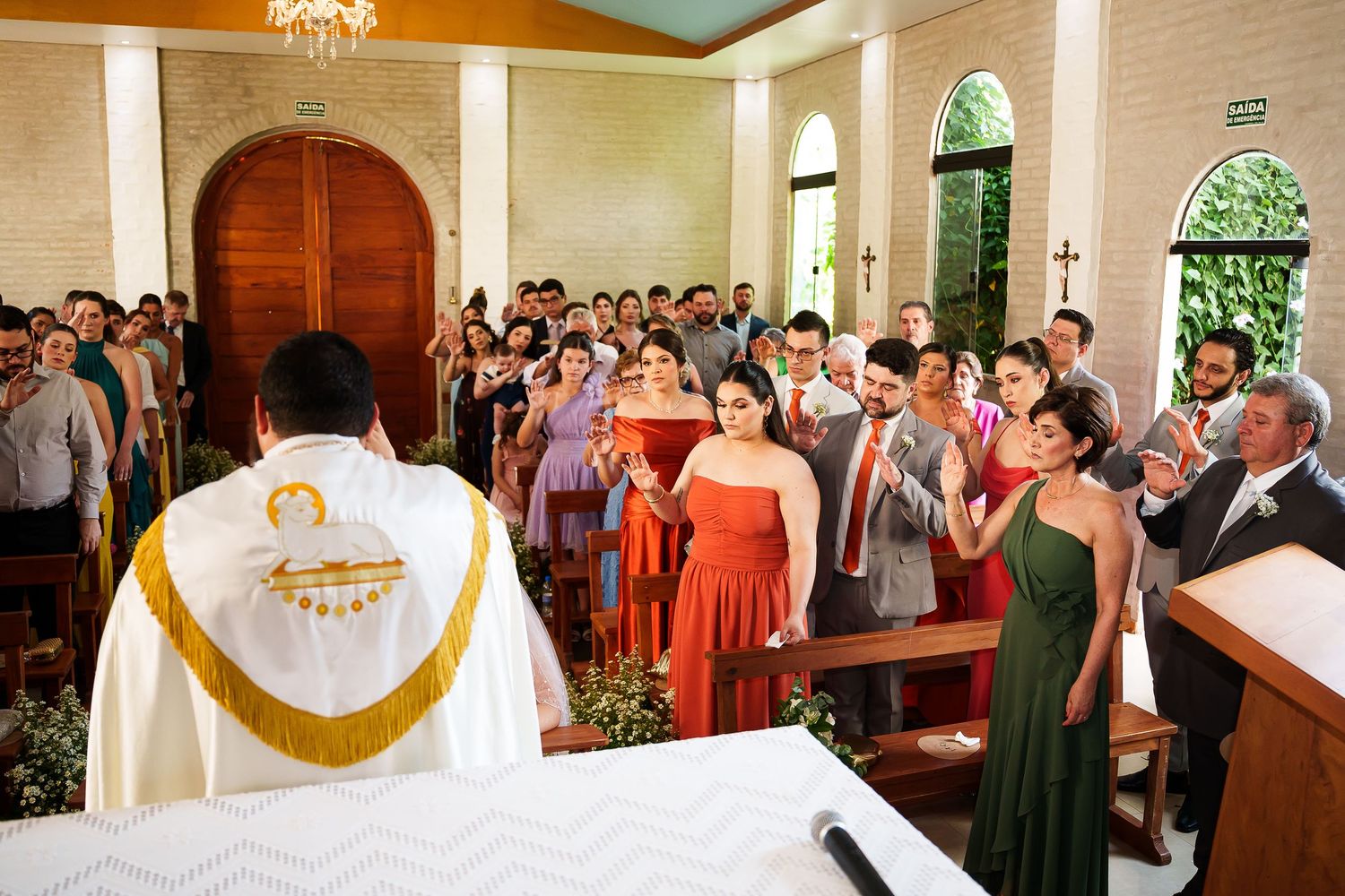 Catholic wedding ceremony in a white chapel with bridesmaids in red dresses and guests standing before a priest at the altar.