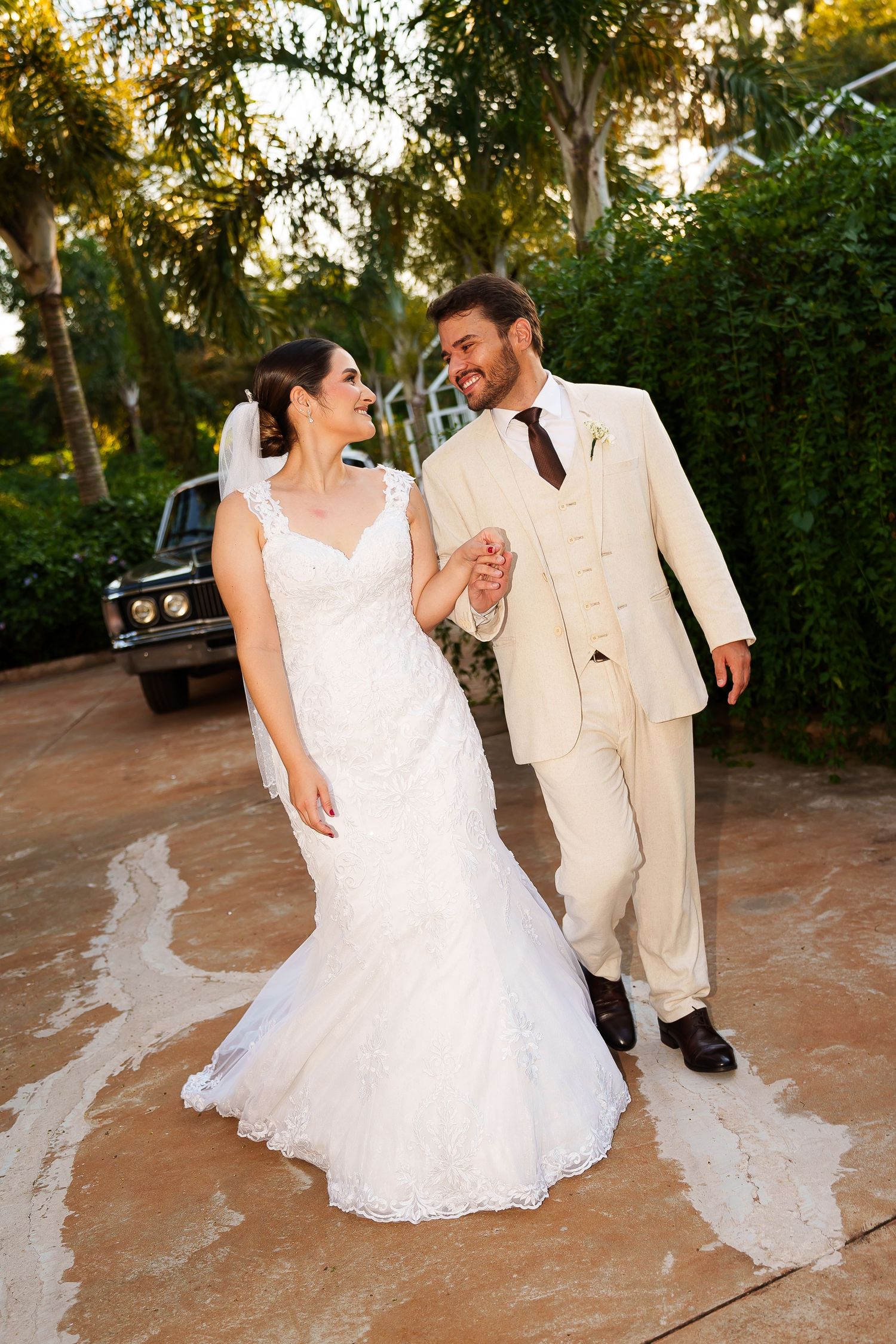 Bride in white lace gown and groom in cream suit hold hands, smiling, walking outdoors near vintage car.