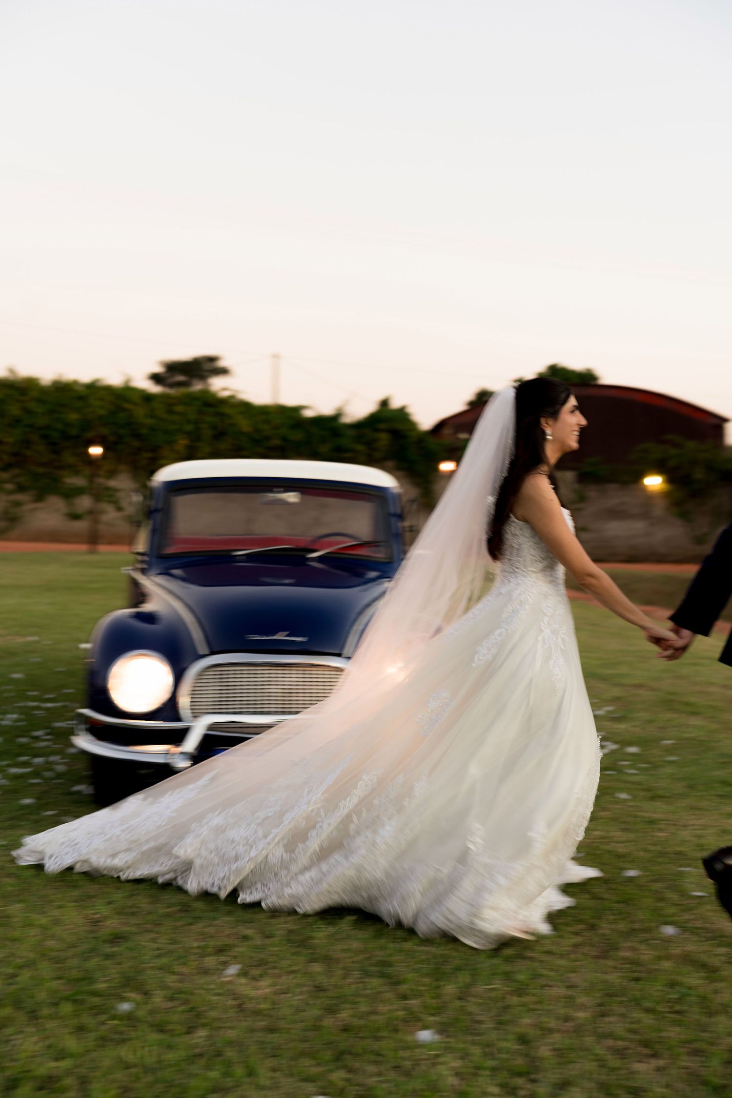 Bride in lace wedding gown with long veil walks hand-in-hand near vintage blue car at dusk.