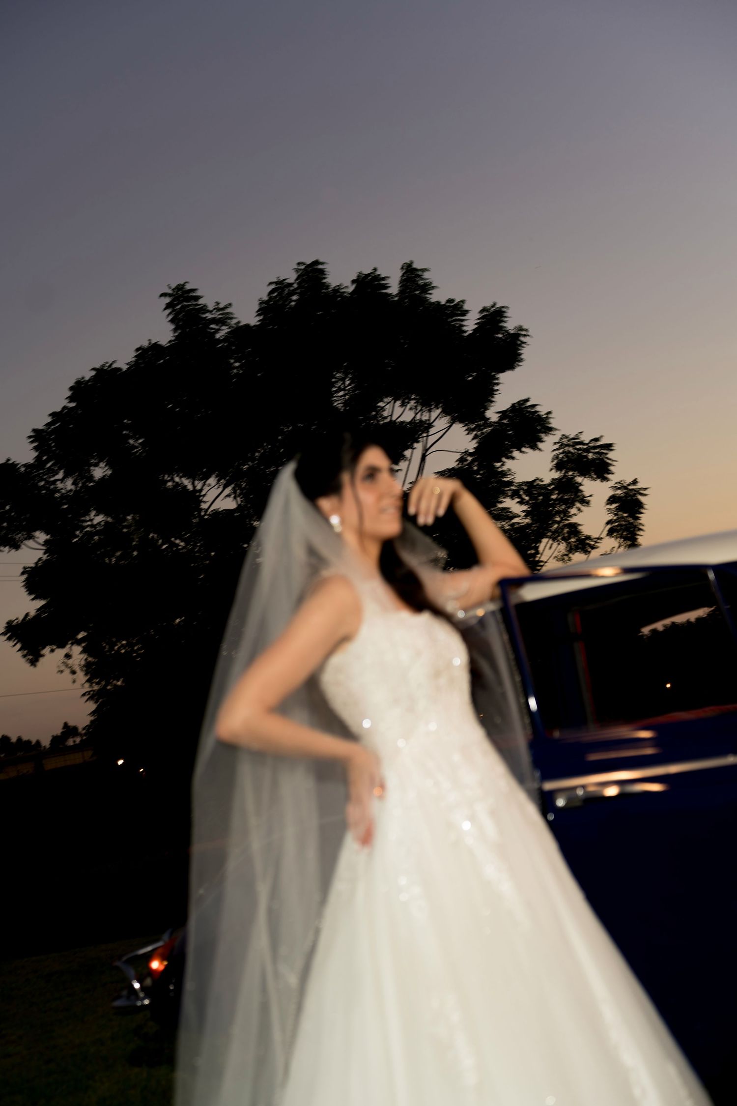 Bride in white wedding gown and veil poses beside a classic car at dusk with trees silhouetted against twilight sky.