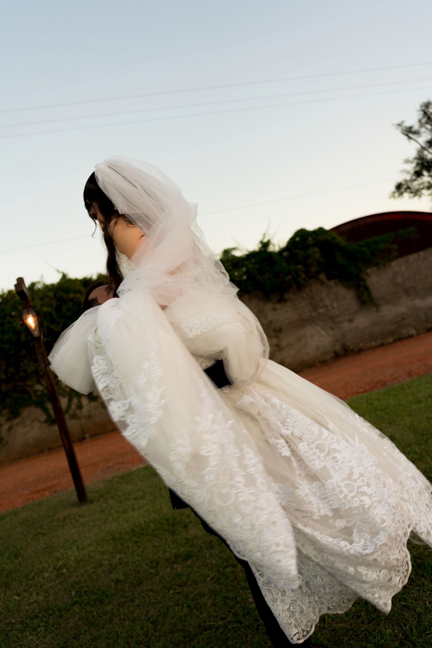 Bride in white lace wedding dress and veil seated outdoors at dusk near lush green vineyard.