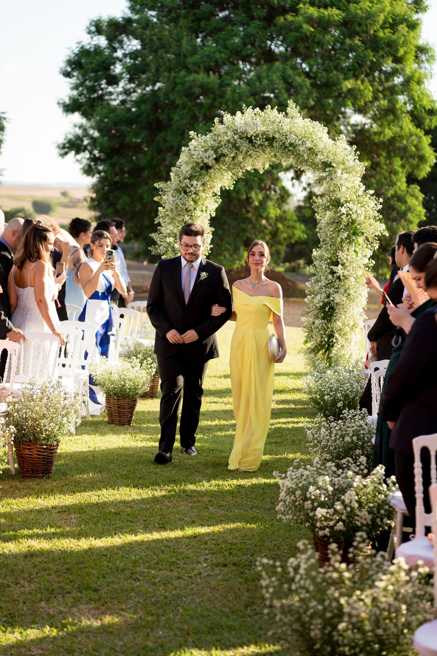 Couple walks down outdoor wedding aisle under floral arch, woman in yellow gown, surrounded by guests on green lawn.