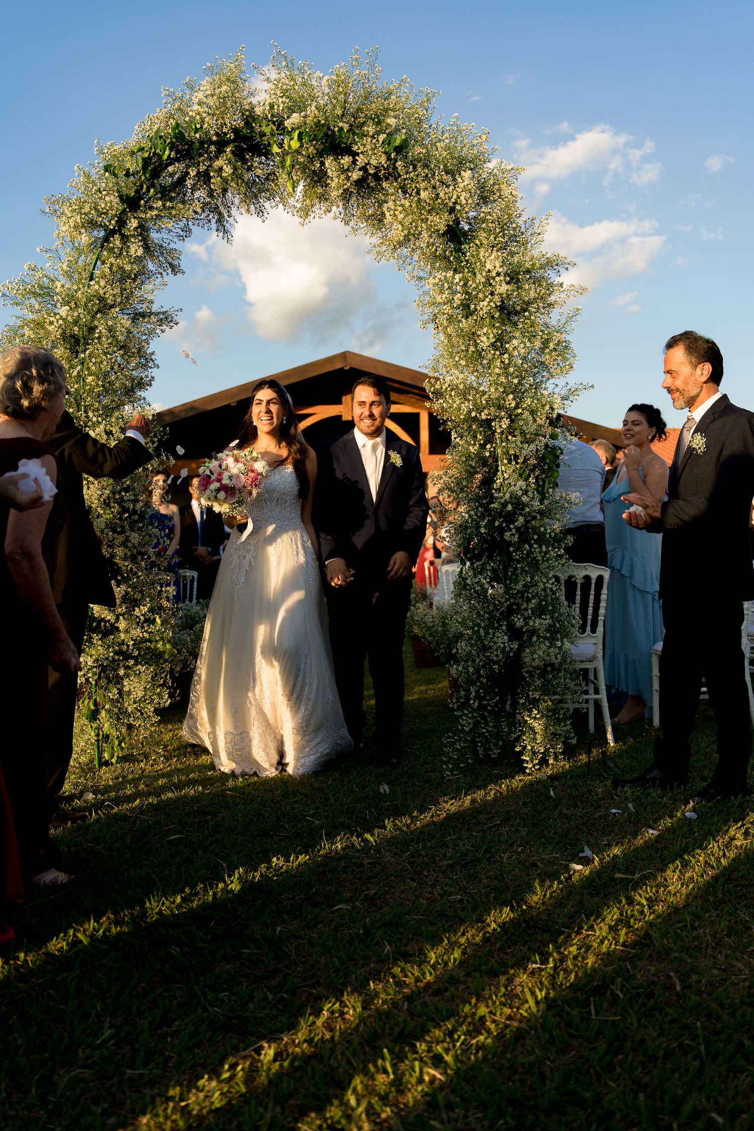 Bride and groom walk under floral arch at outdoor wedding ceremony, guests cheering on sunny day.
