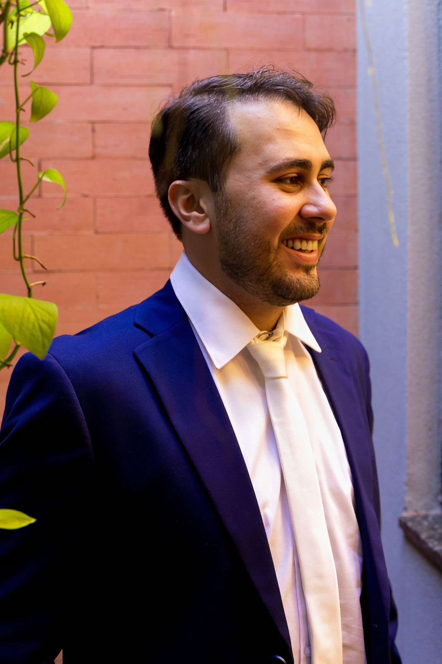 Smiling man in navy blue suit and gold tie standing outdoors near brick wall with green vines.