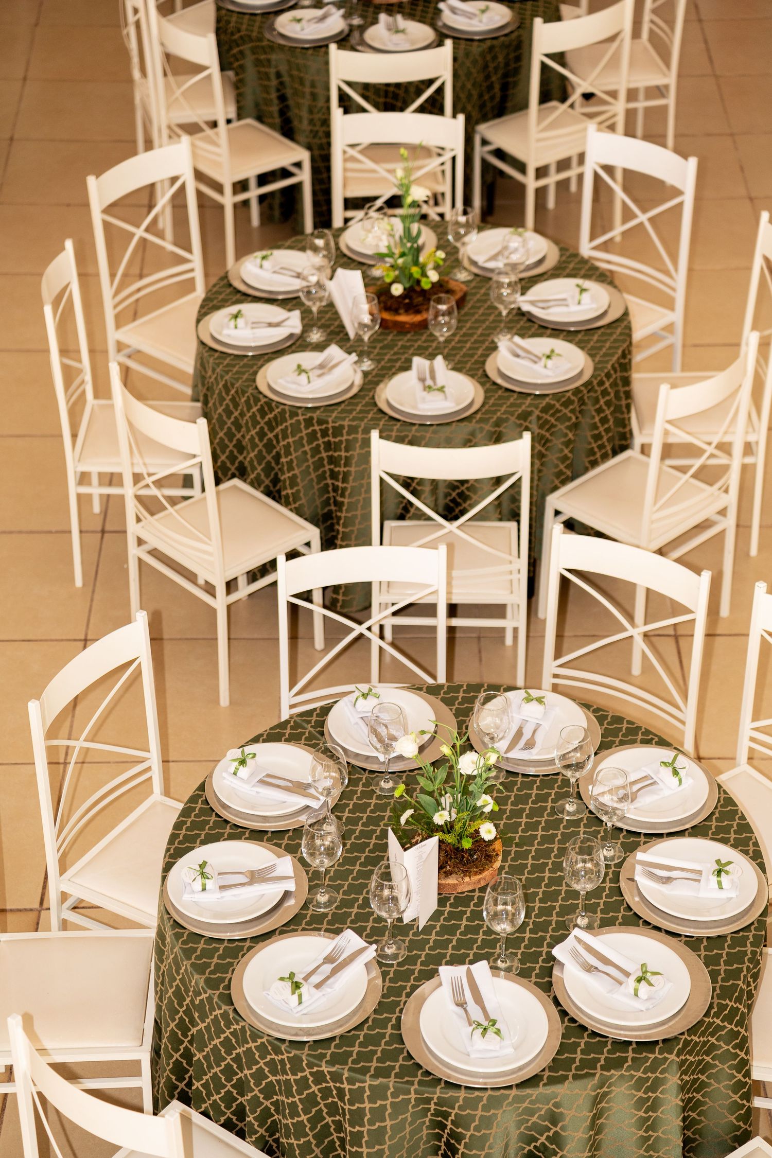 Elegant event venue with round tables draped in green mosaic linens, white cross-back chairs, and floral centerpieces.