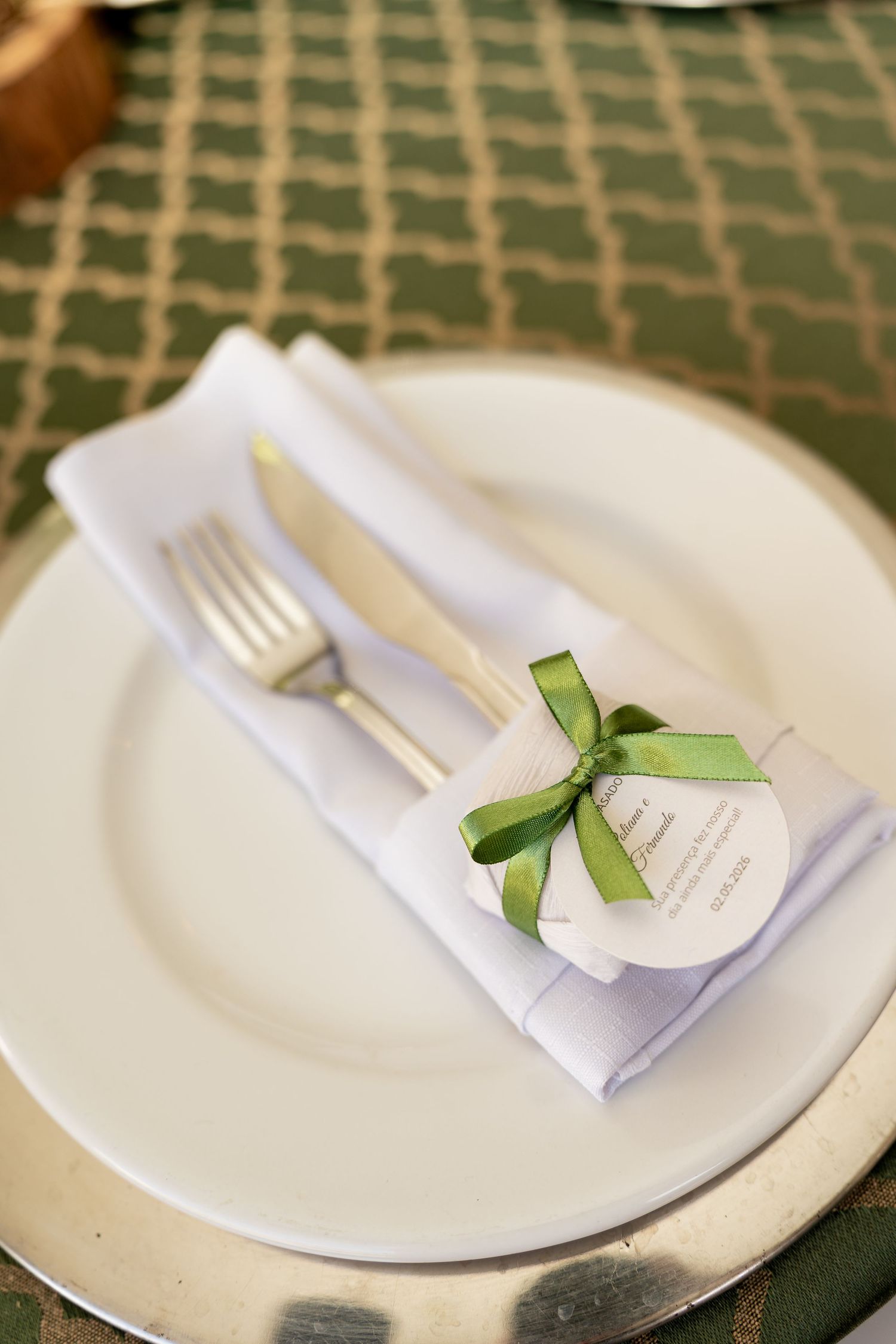 Elegant place setting with white plate, silver cutlery, folded napkin, and green ribbon tag on green patterned tablecloth.
