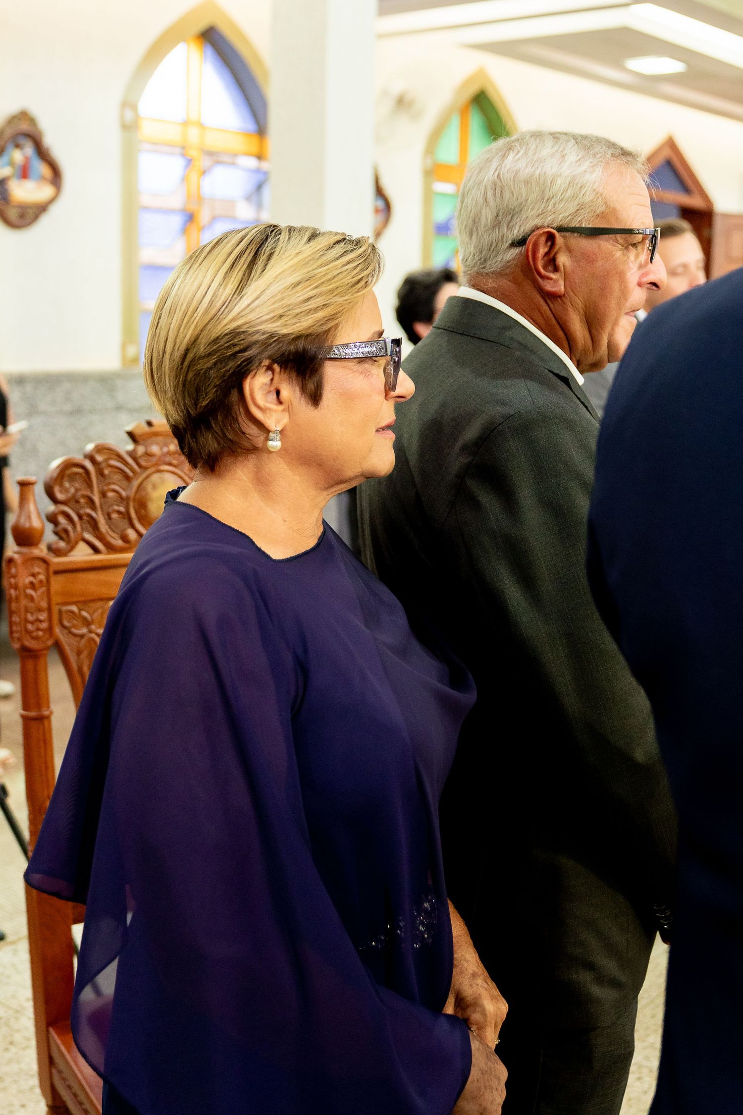 Elegant couple standing in a church during a formal ceremony, woman in purple blouse with short blonde hair and glasses.