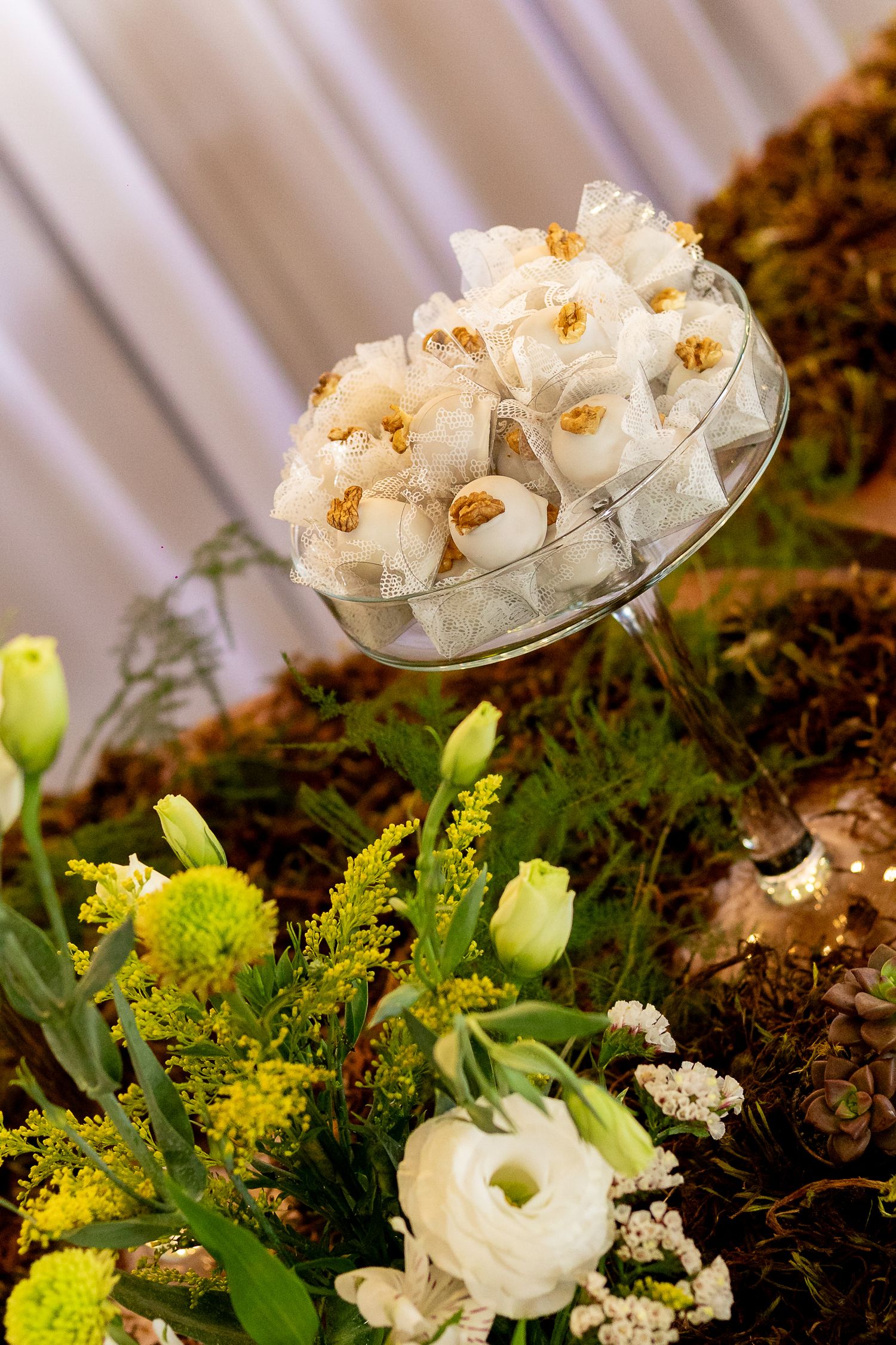 Elegant wedding favor display with white tulle bags in glass bowl surrounded by white ranunculus and yellow flowers.