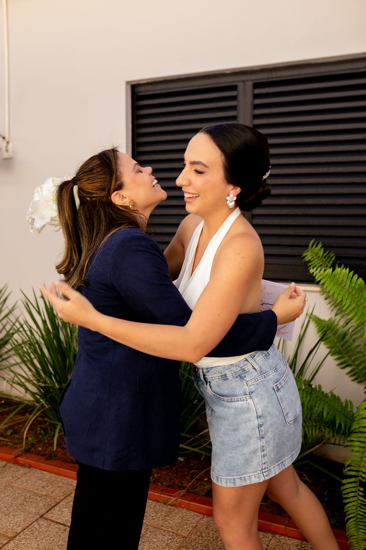 Two women sharing a warm embrace and laughing together outdoors, one in a navy blazer and one in a white top and denim skirt.