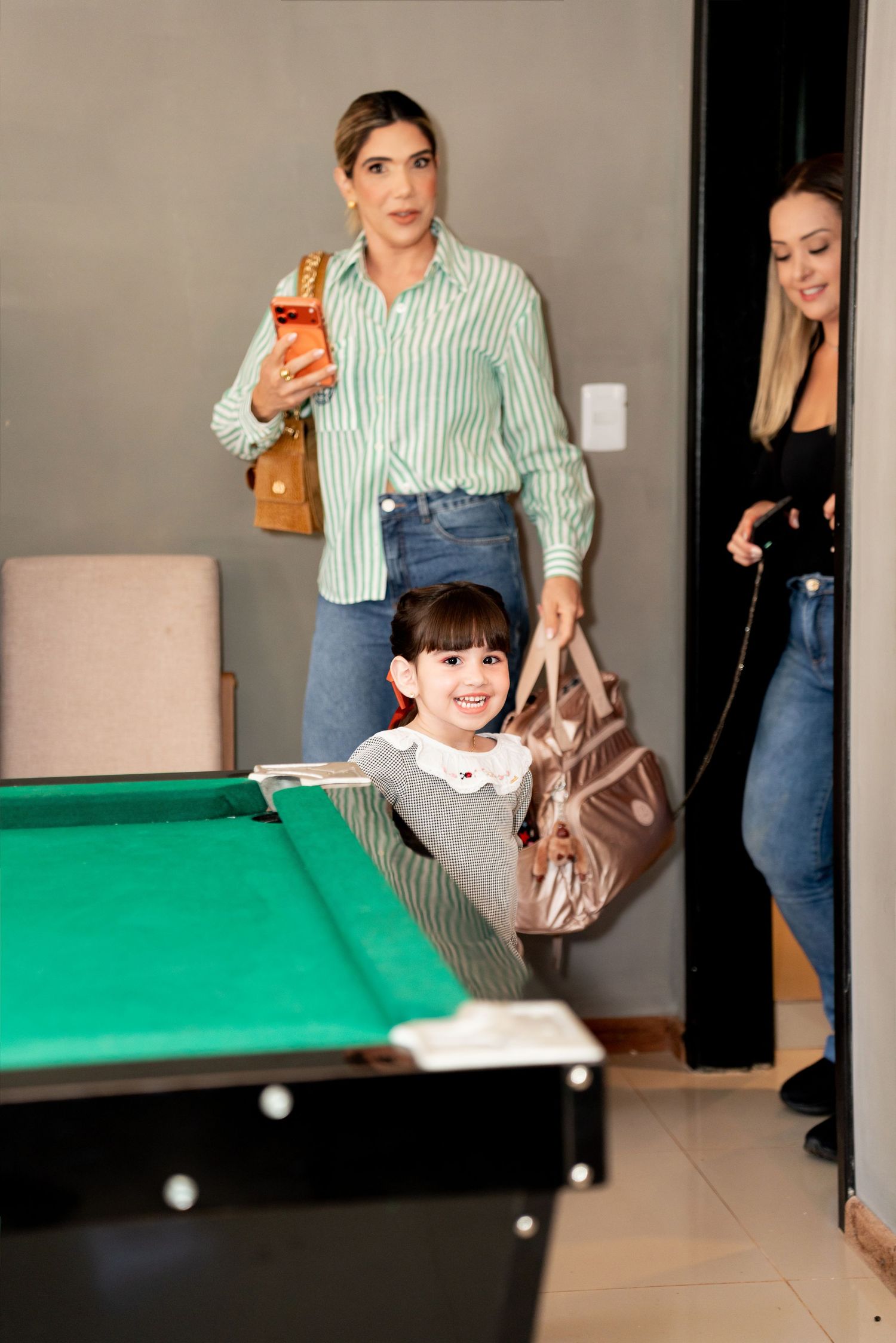 Woman in striped shirt holding child and bag near pool table, with another woman entering through doorway.