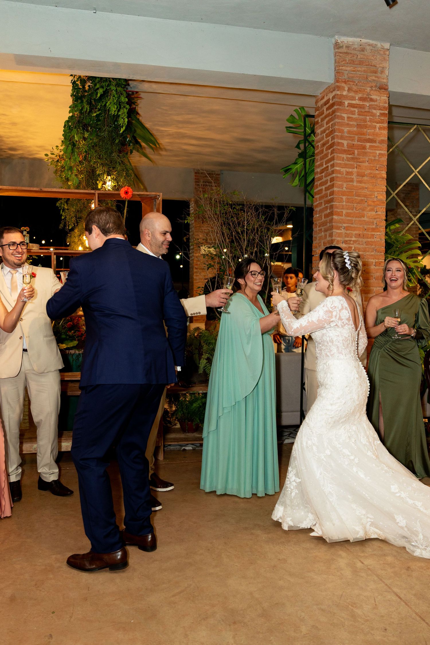 Bride in white lace gown and groom in navy suit share a dance with guests in elegant green dresses at wedding reception.