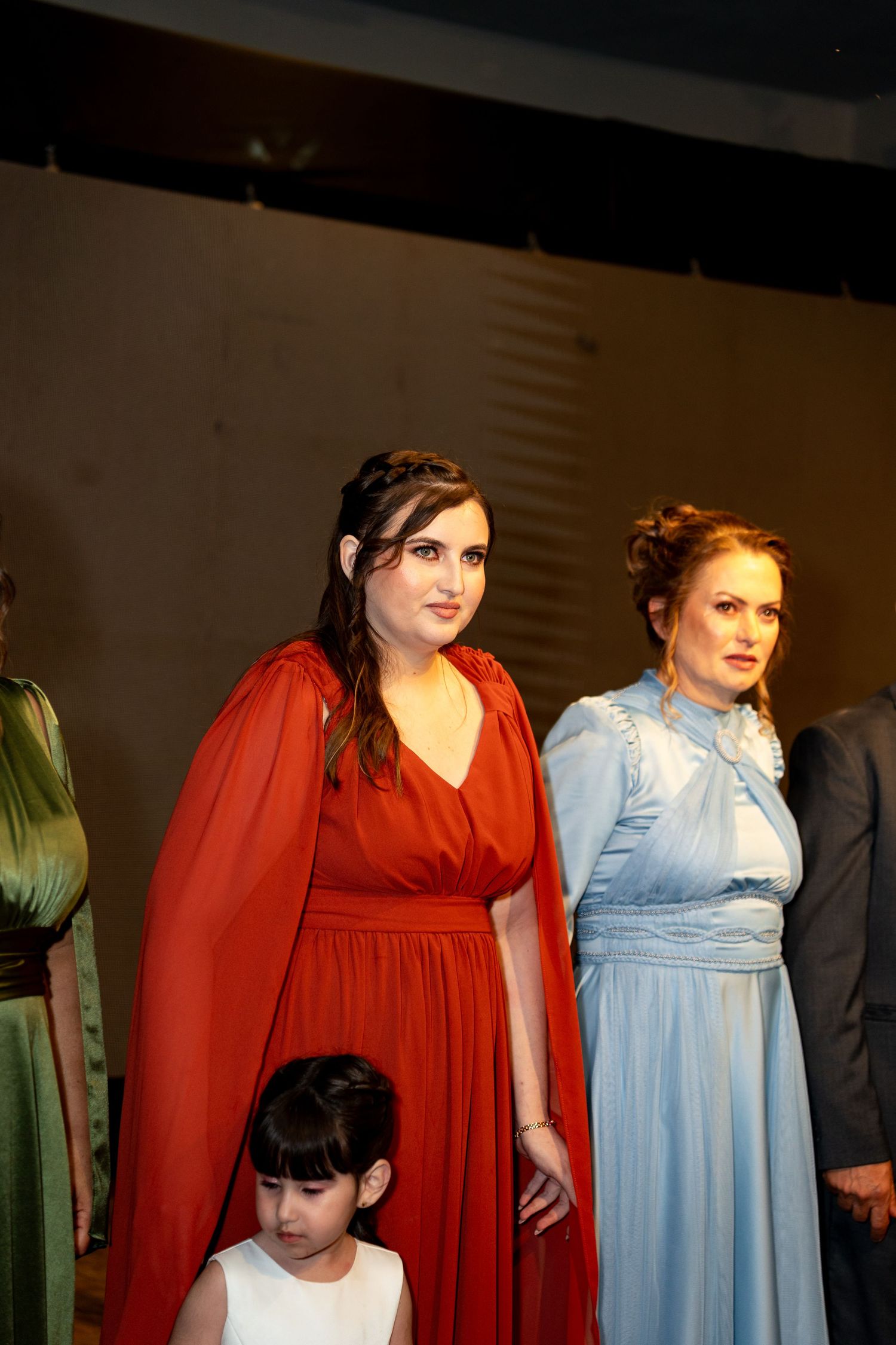 Three women in elegant gowns, red, blue, and green, stand on stage during a formal theatrical performance.