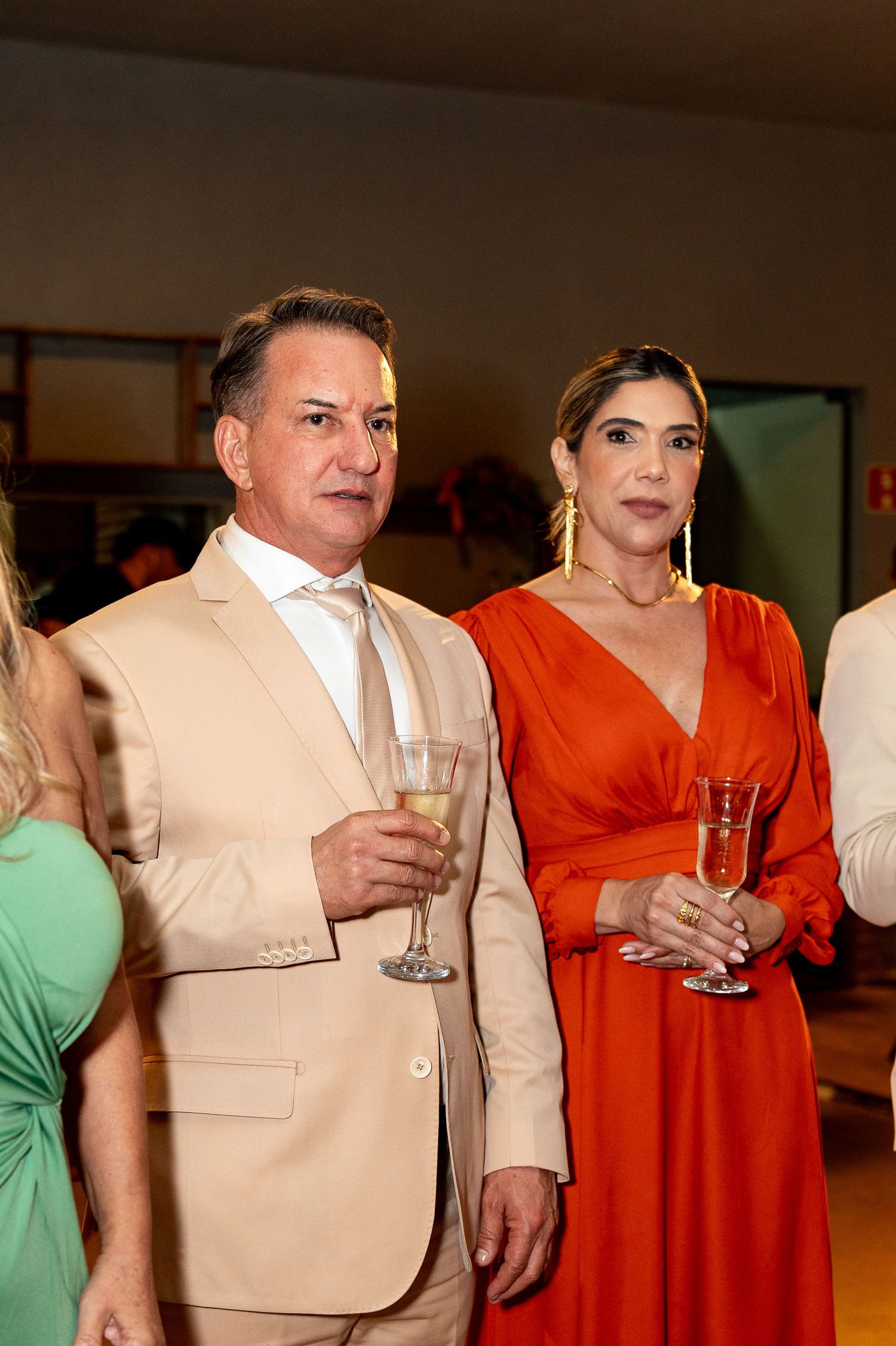 Elegant couple at formal event, man in beige suit with champagne, woman in red dress with gold earrings.