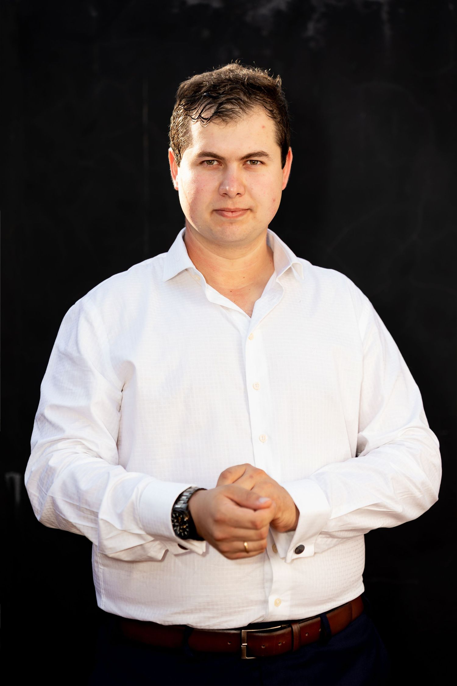 Professional man in white dress shirt with watch, posing confidently against dark background.