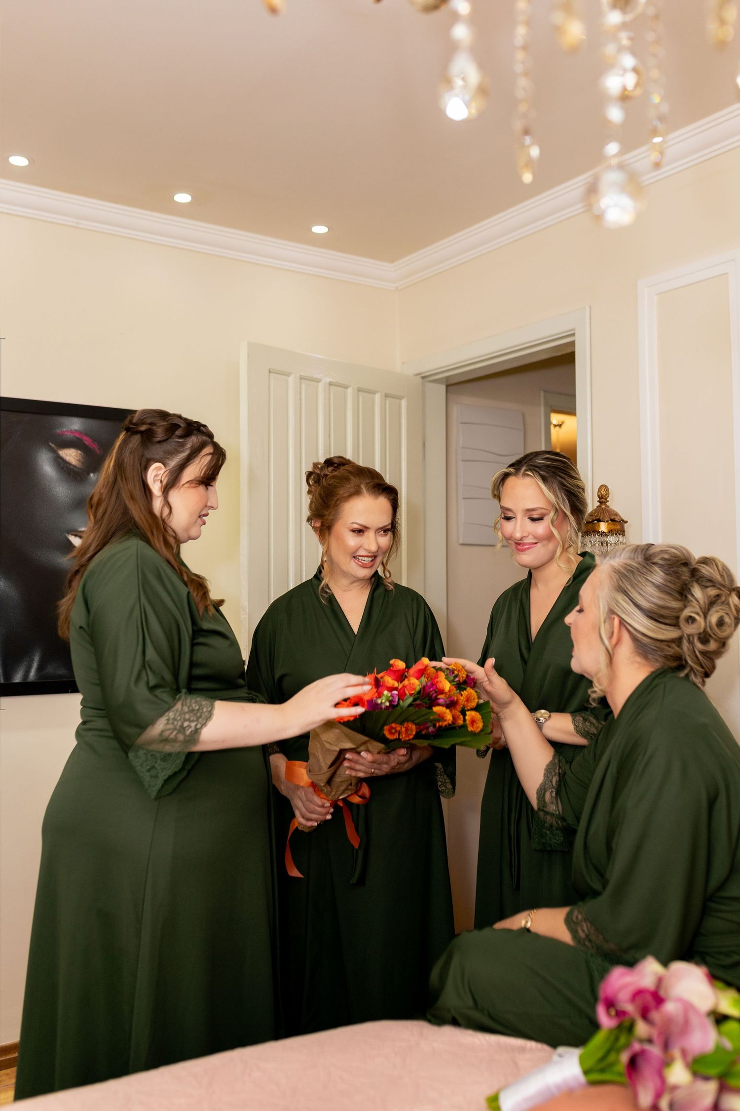Four bridesmaids in green dresses admire a colorful bouquet of orange and red flowers indoors.