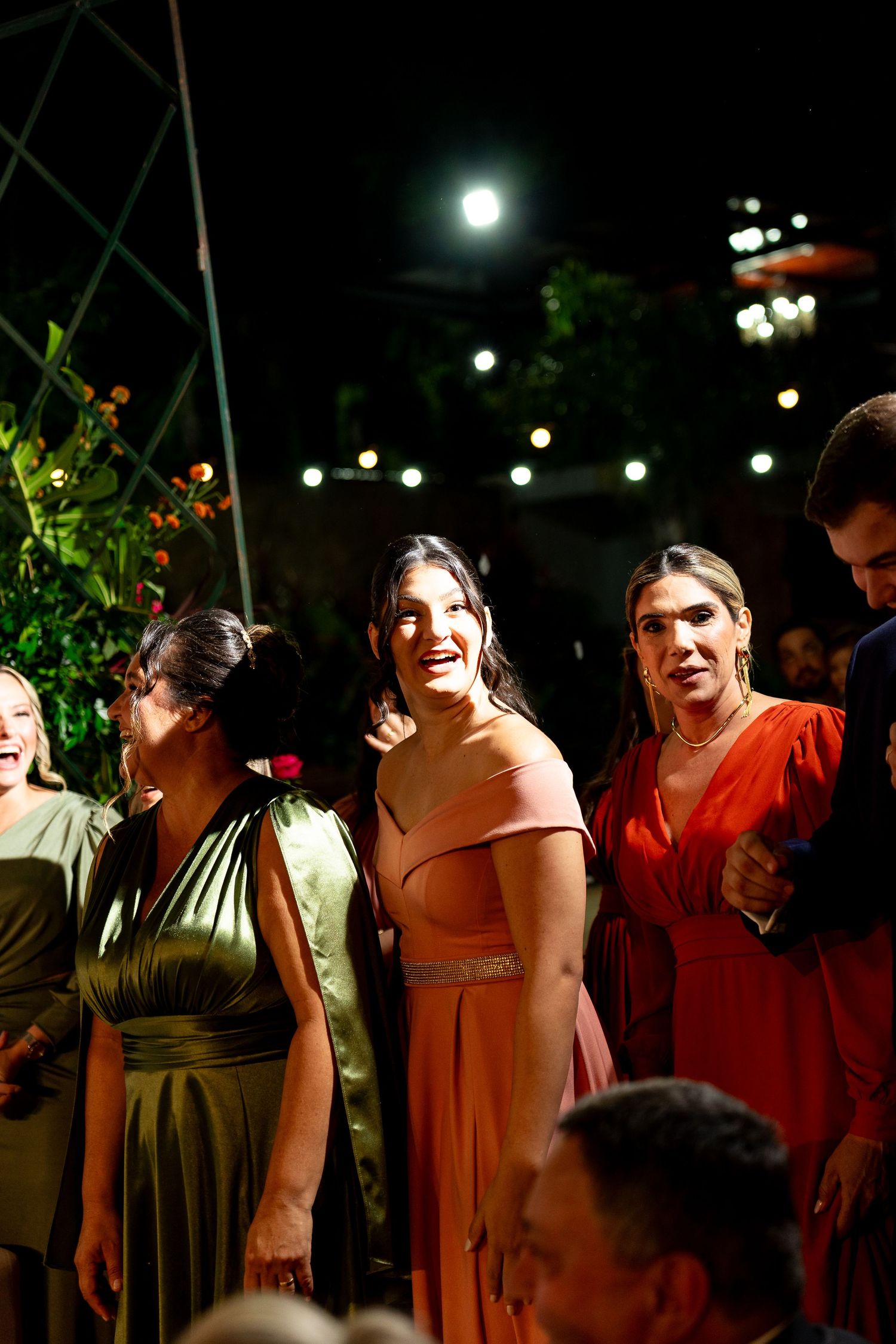 Women in elegant colorful gowns smiling at an outdoor evening event with string lights in background.