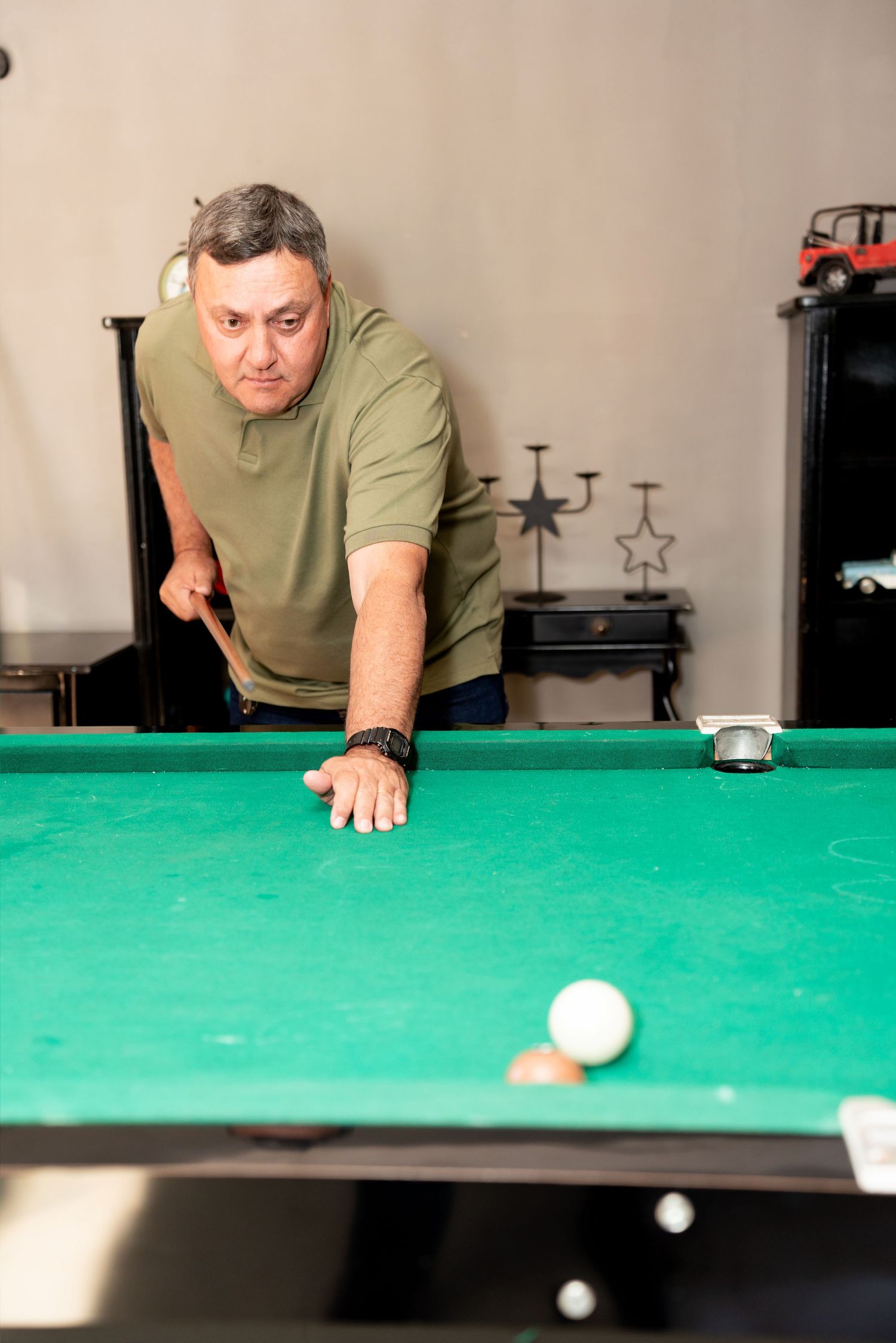 Middle-aged man in green shirt leaning over a pool table, aiming his cue stick at billiard balls.