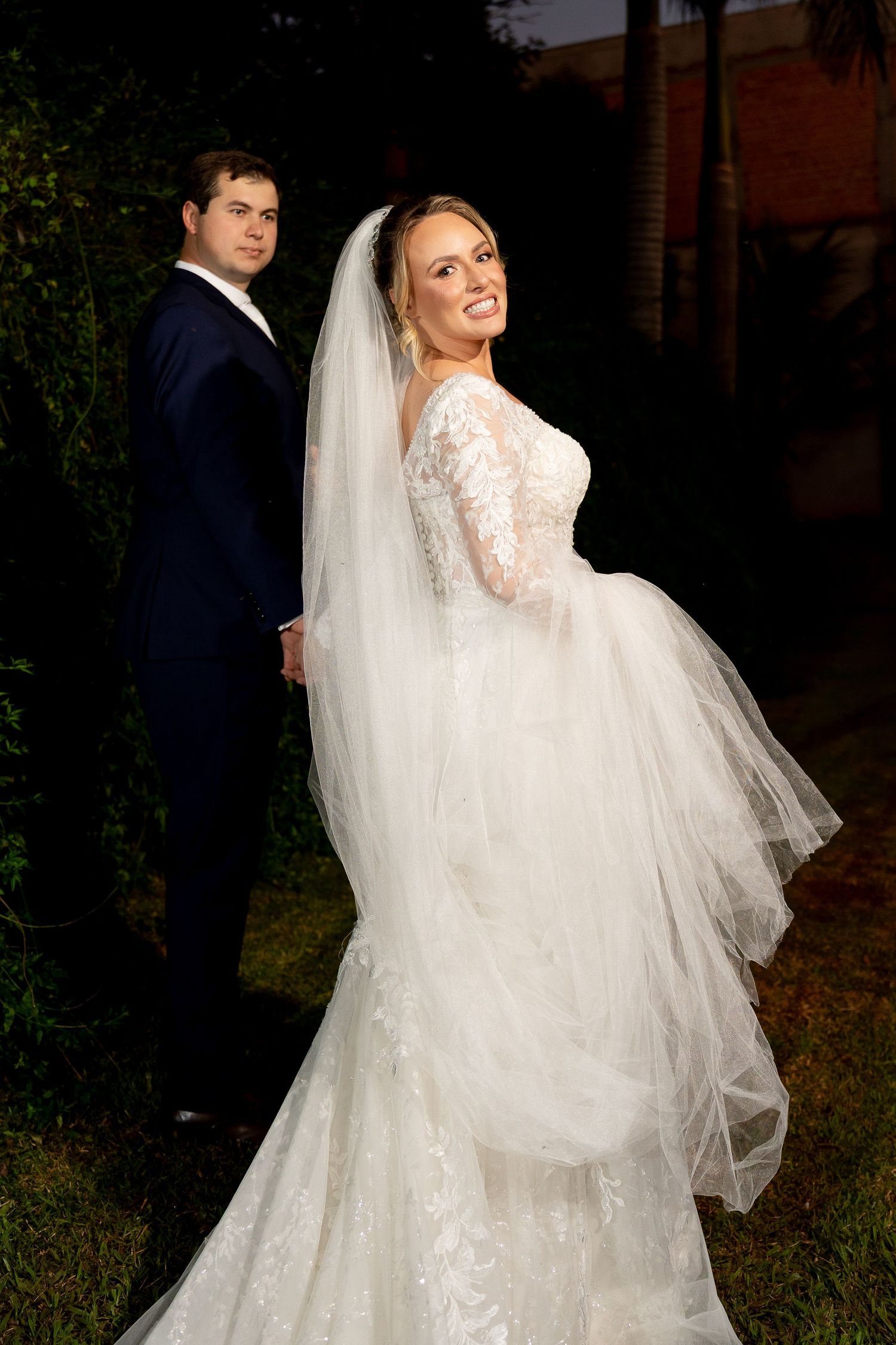 Bride in lace long-sleeve wedding gown with veil smiles over shoulder beside groom in navy suit outdoors at night.