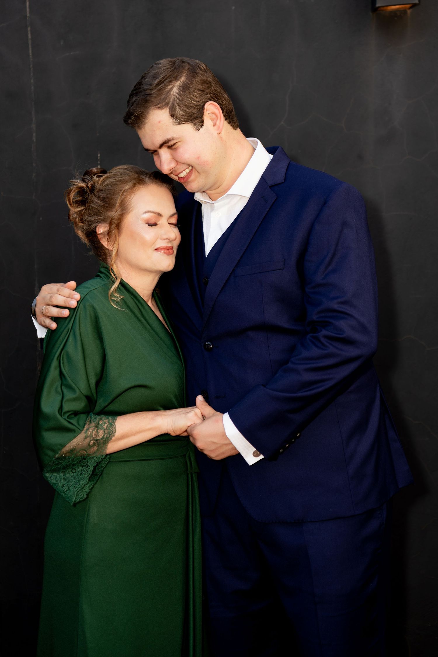 Couple in elegant attire, woman in green dress and man in navy suit, sharing a tender embrace against dark background.