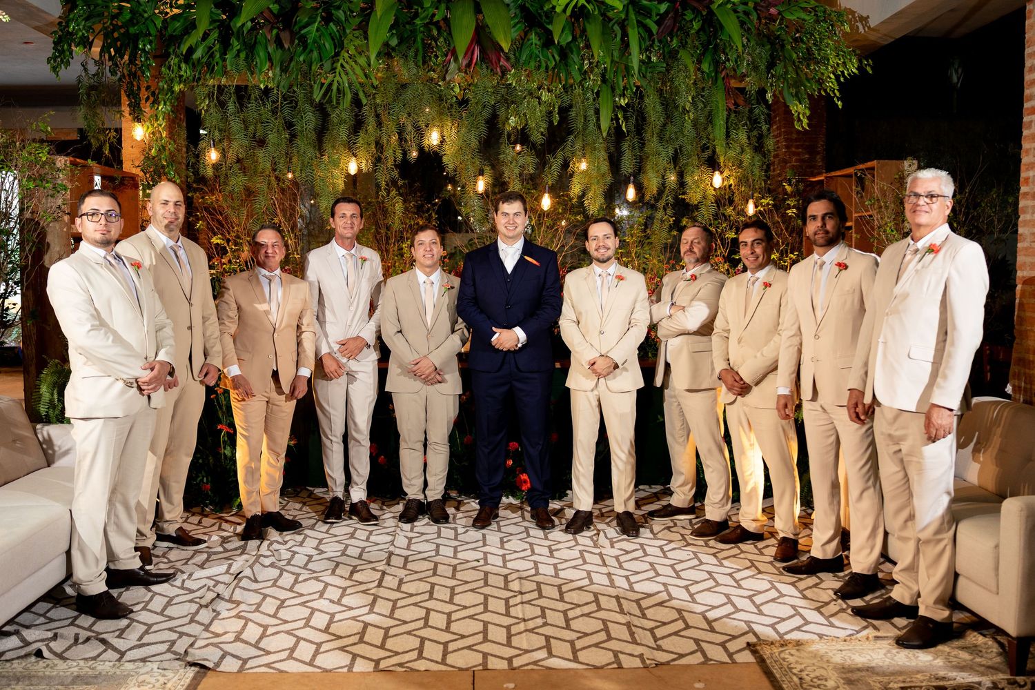 Groom in navy suit stands with groomsmen in tan suits, posing indoors with lush greenery backdrop.