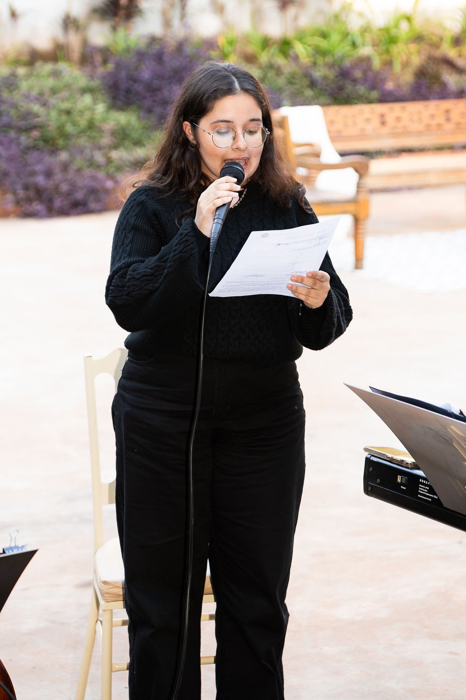 Young woman in black outfit reads from paper at microphone stand outdoors at formal event.