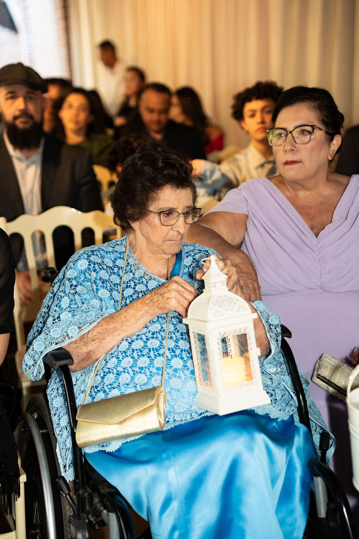 Elderly woman in blue dress holding lantern in wheelchair at formal event, accompanied by woman in lavender.
