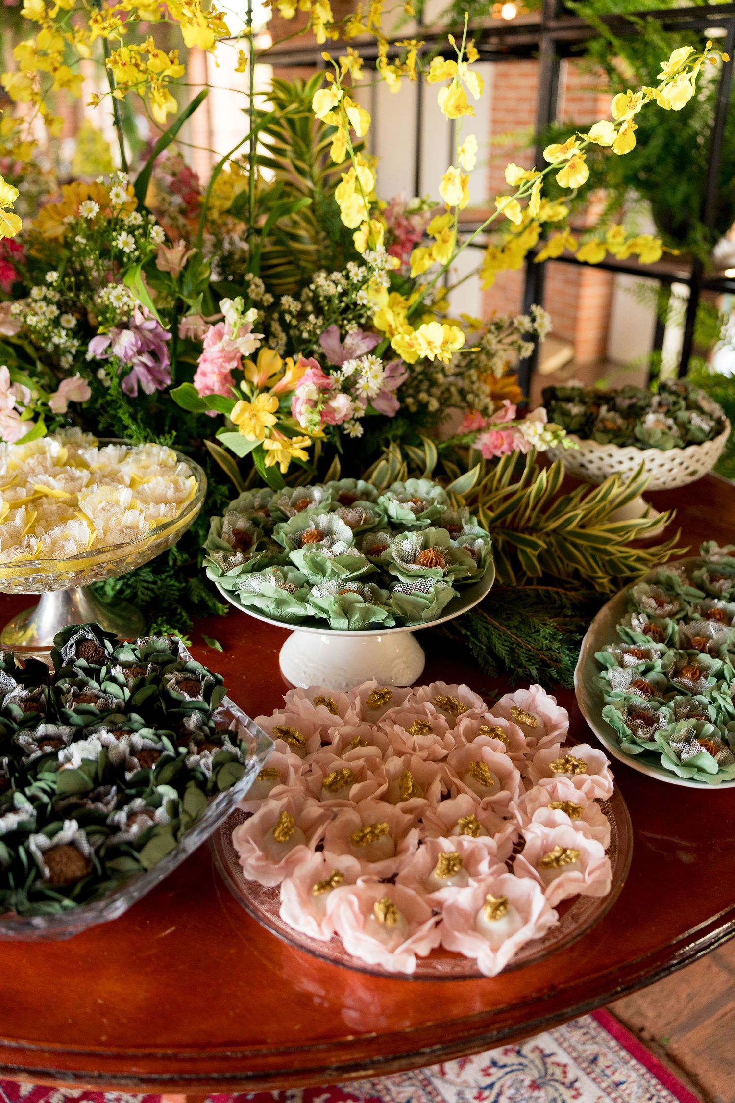 Elegant dessert table with flower-shaped sweets and lush floral arrangements including yellow orchids at a festive event.