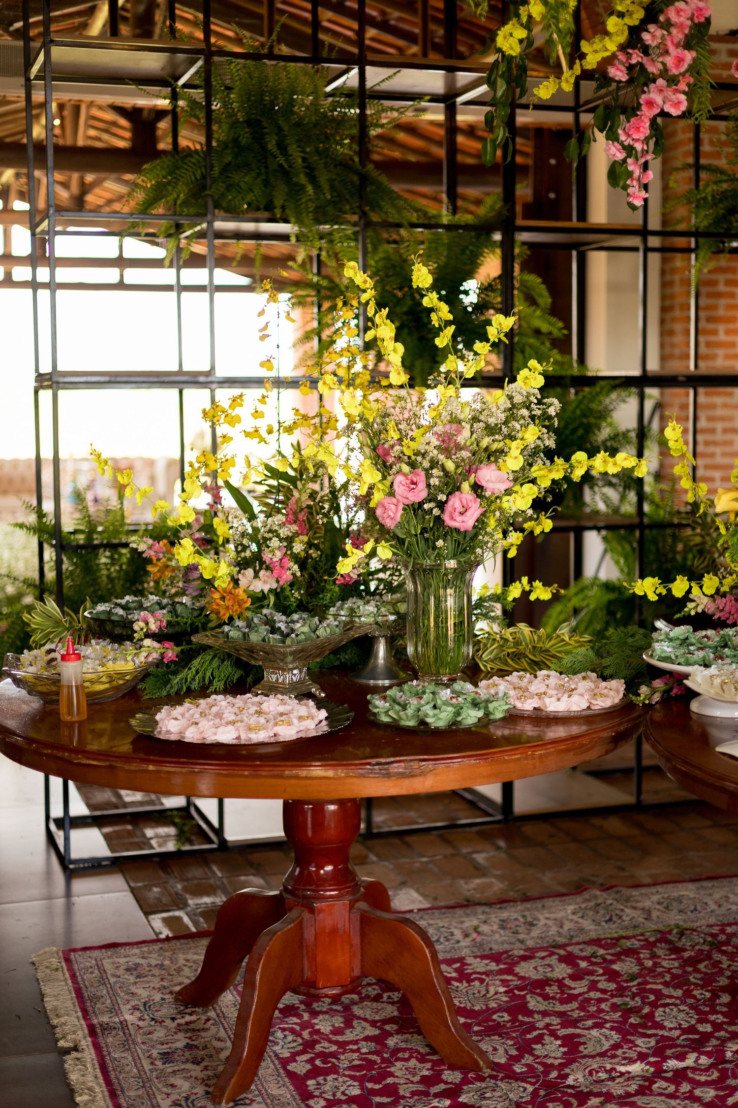 Elegant wooden round table with floral arrangements of yellow and pink flowers, surrounded by lush greenery in a rustic venue.