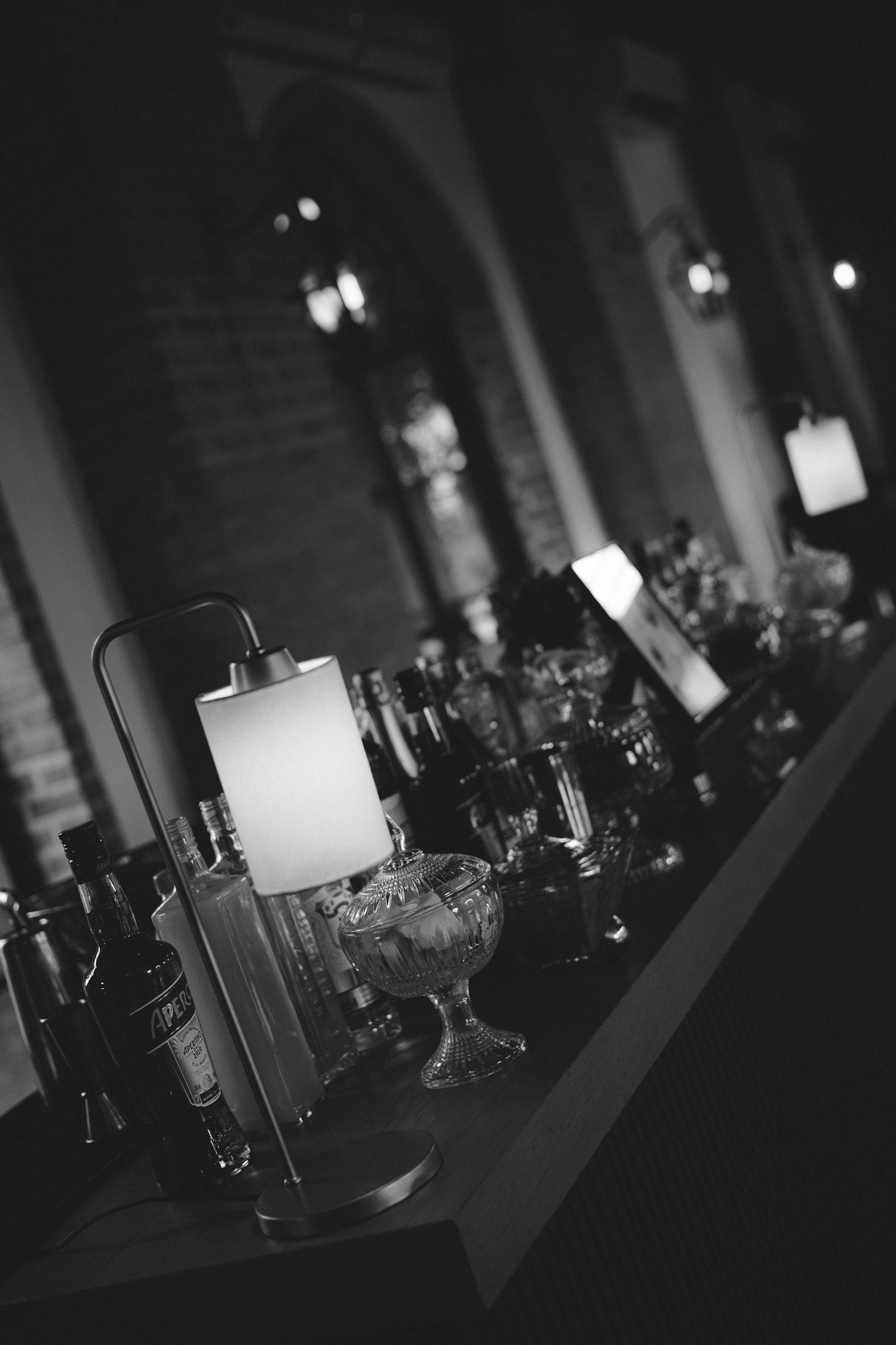 Black and white photo of elegant table lamp and glassware on a bar counter in a dimly lit venue.