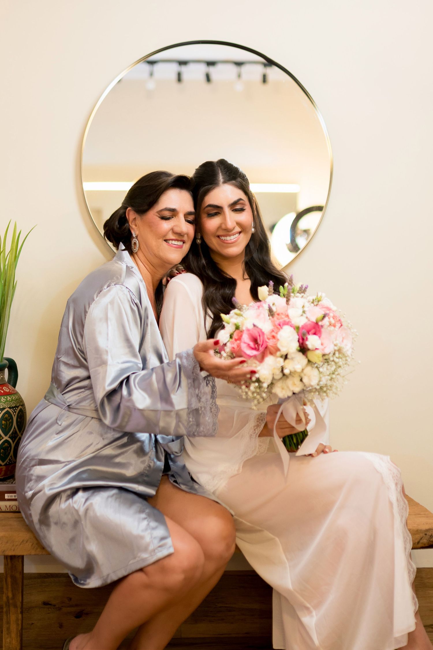 Two women in satin robes sit together holding a pink bridal bouquet in front of a round mirror.