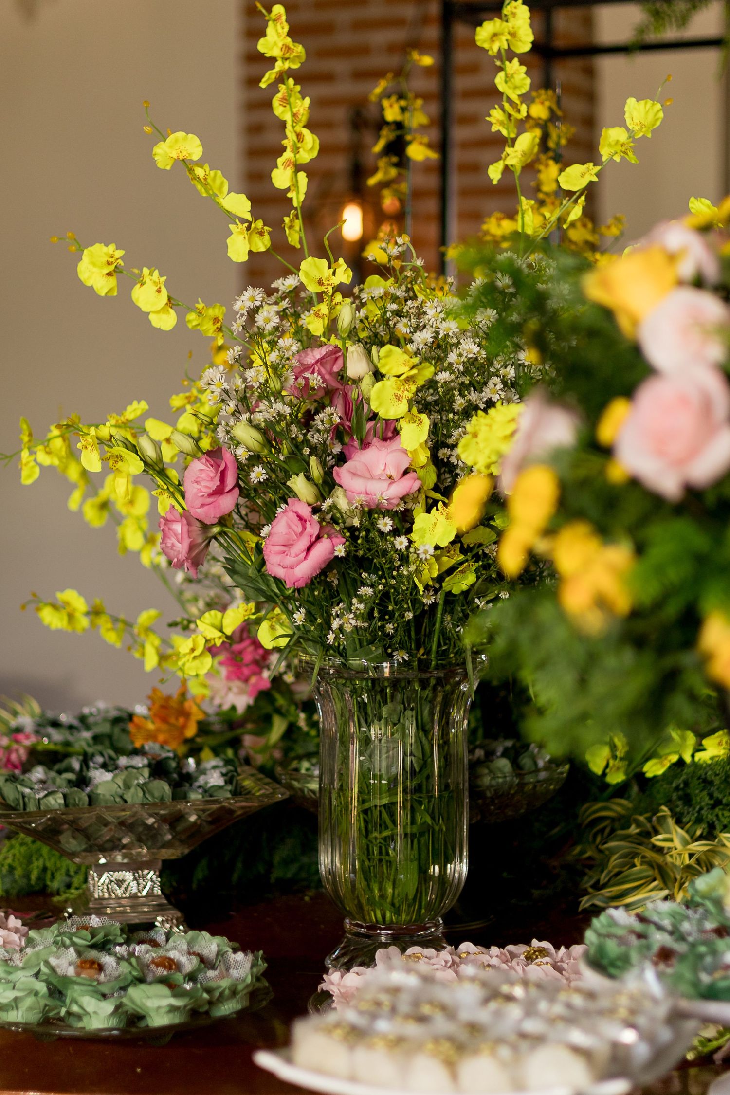 Glass vase with pink roses and yellow flowers on a table with green desserts at an elegant event.