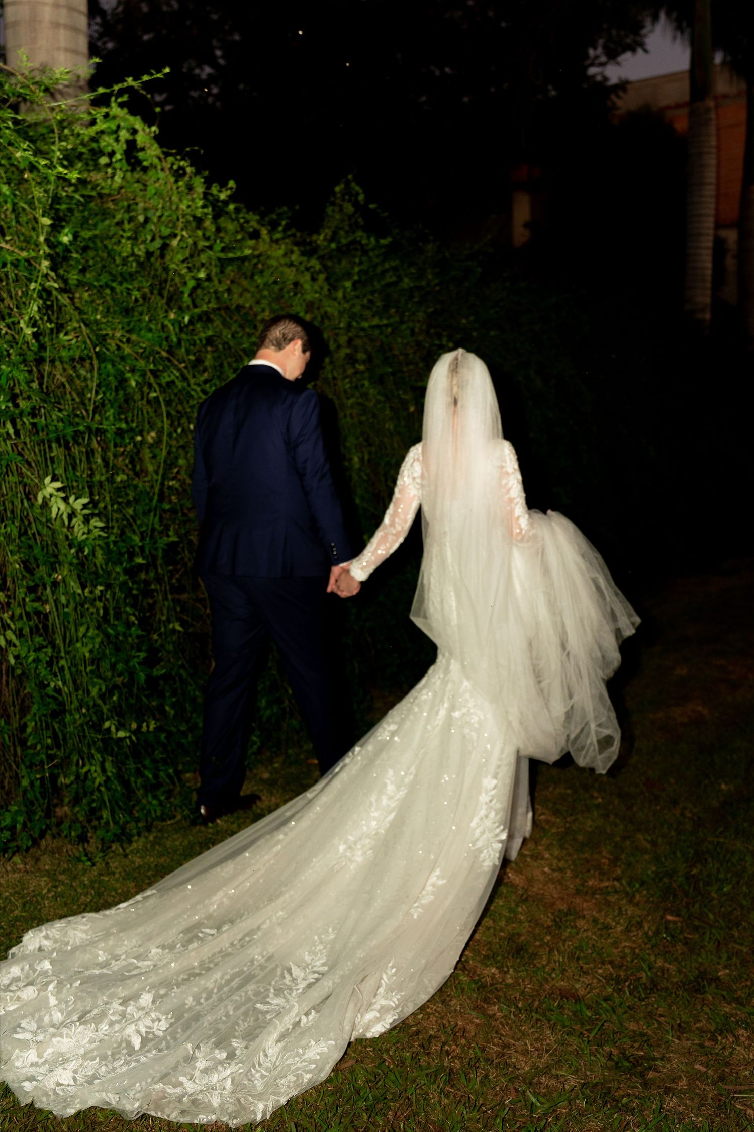 Bride and groom holding hands walking at night, elegant lace wedding gown with long train and veil.