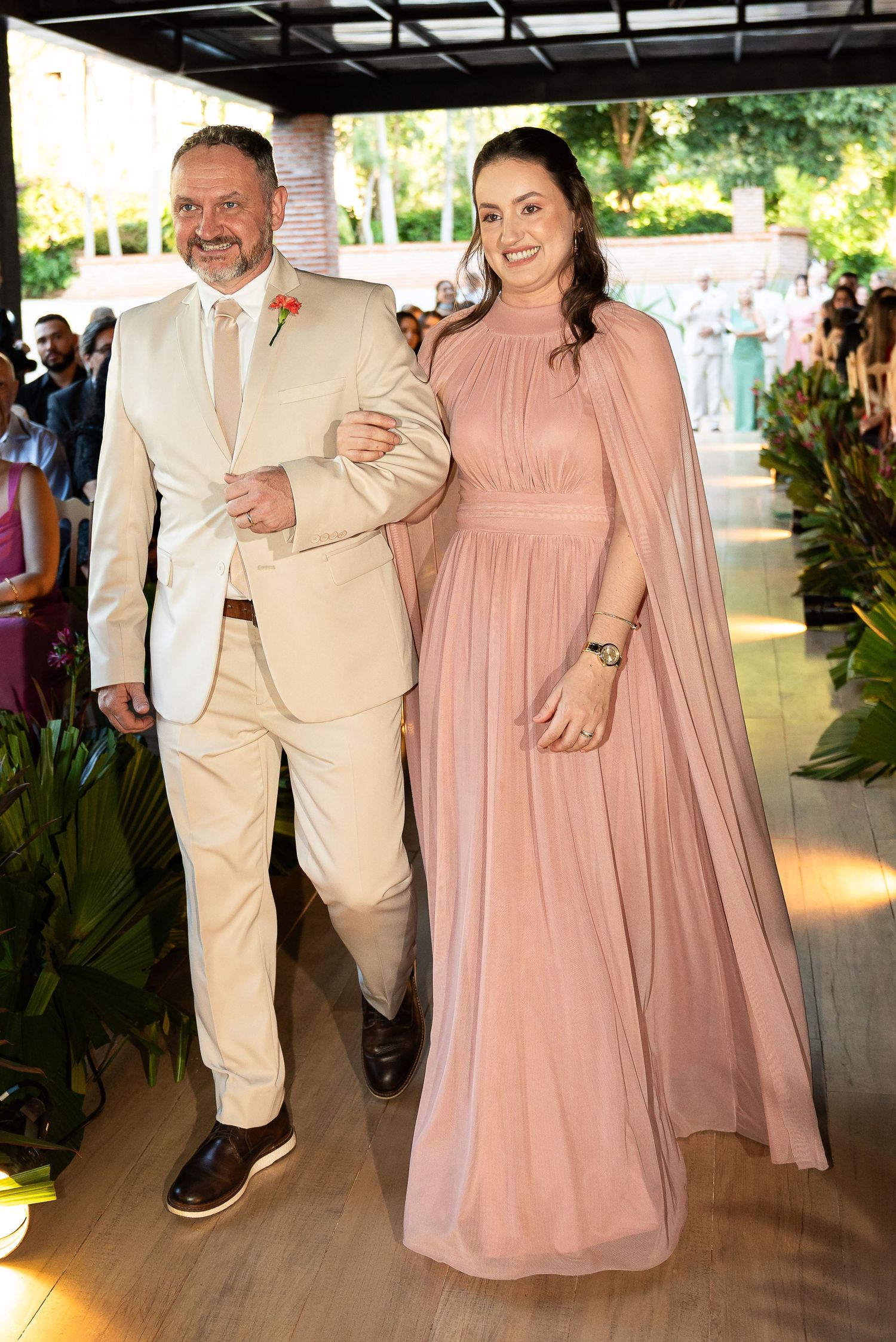 Elegant couple walking at a wedding, man in beige suit, woman in flowing dusty rose cape gown.
