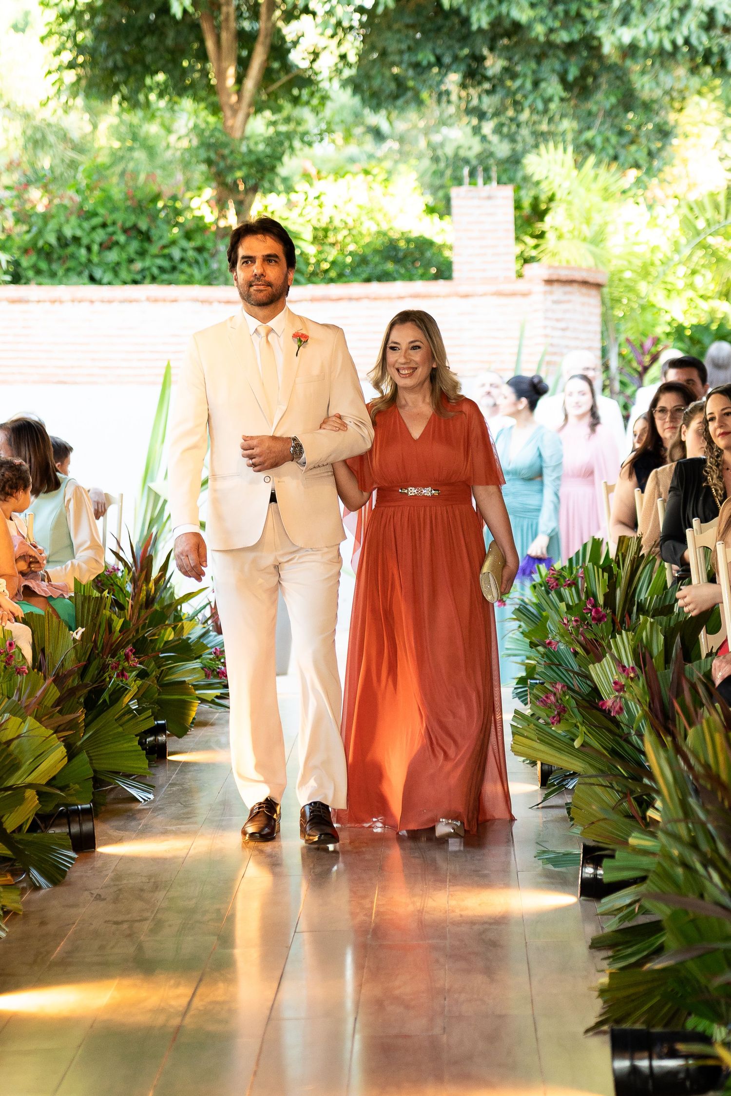 A man in a cream suit escorts a woman in a rust-orange gown down a floral-lined wedding aisle outdoors.