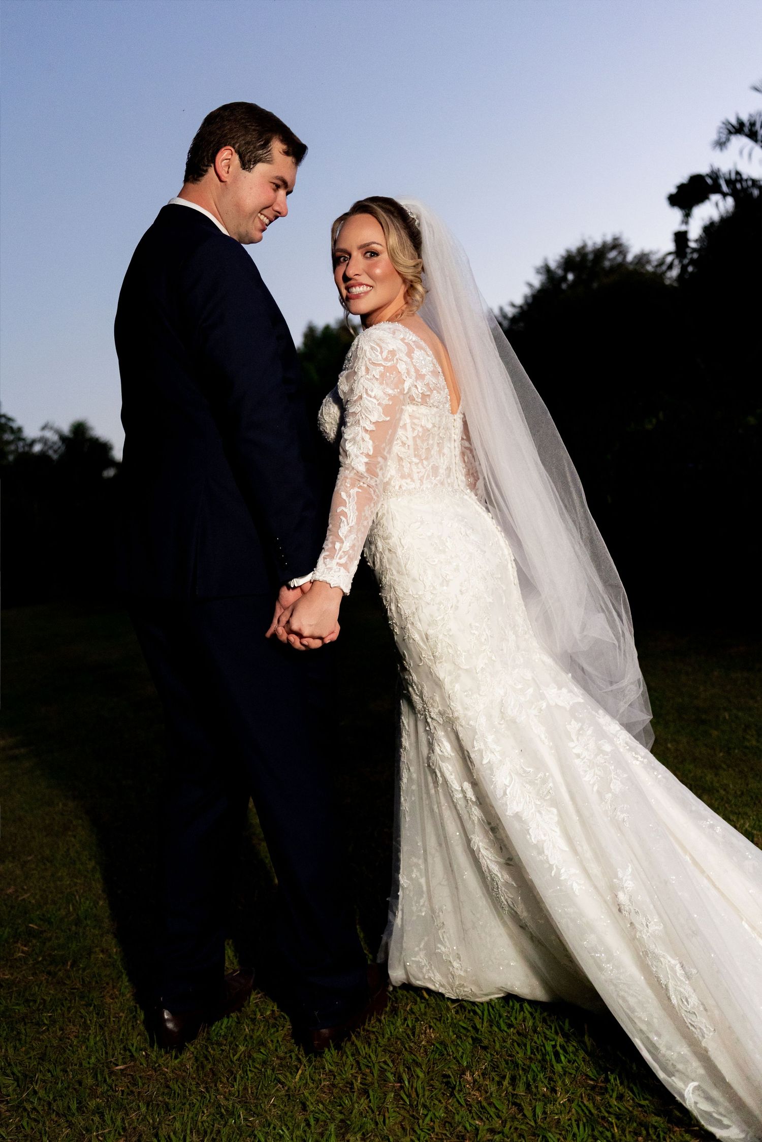 Bride in lace mermaid gown with veil holds hands with groom in navy tuxedo outdoors at golden hour.
