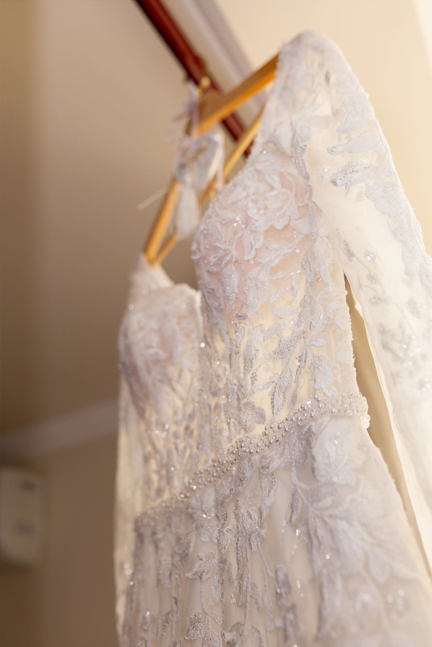 Elegant white lace wedding dress with long sleeves and beaded waist detail hanging on a wooden hanger.