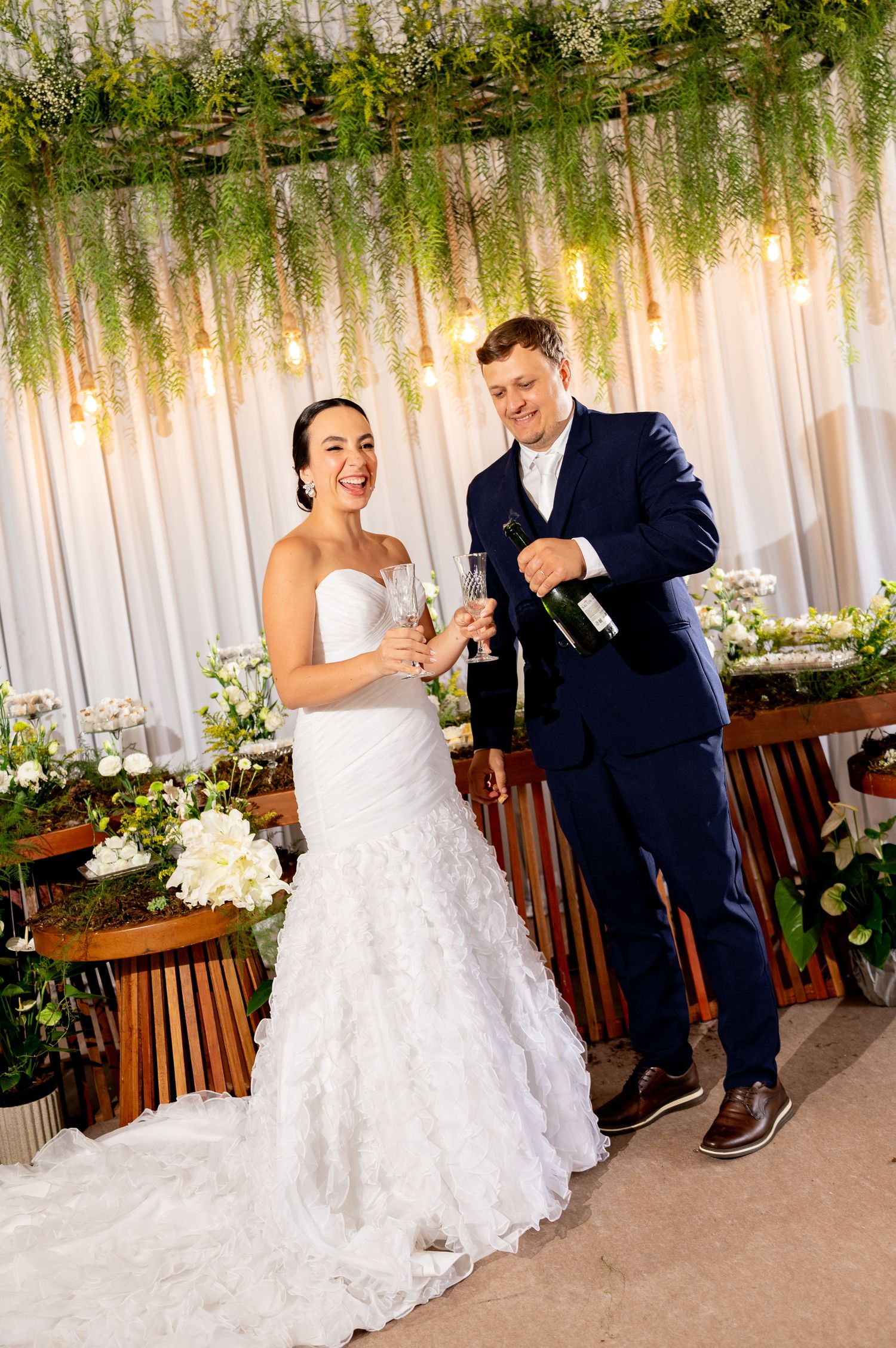 Bride in white gown and groom in navy suit toasting with champagne at elegant wedding reception.