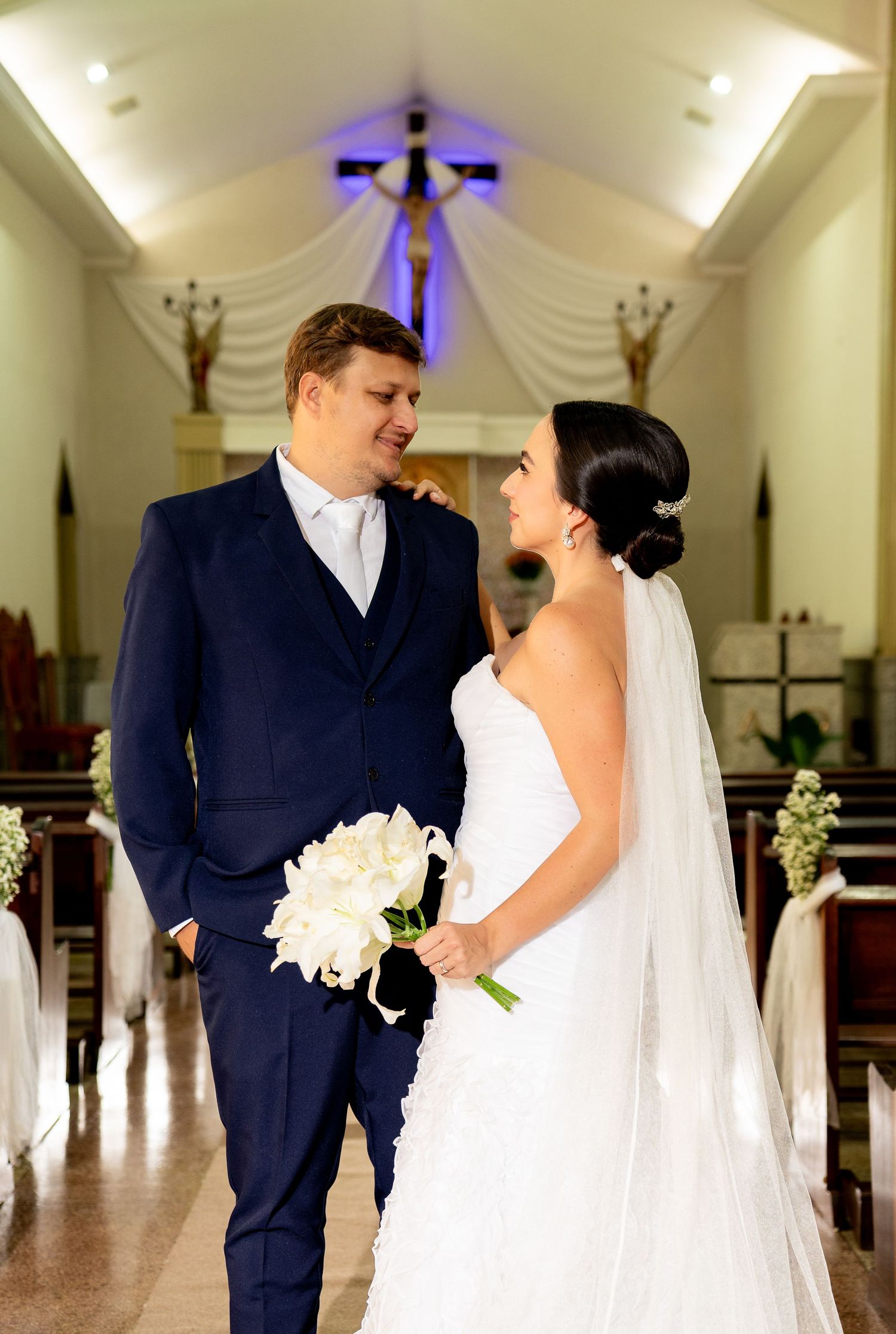 Bride and groom share a loving glance in a church aisle, she holds white peonies in a lace gown with veil.