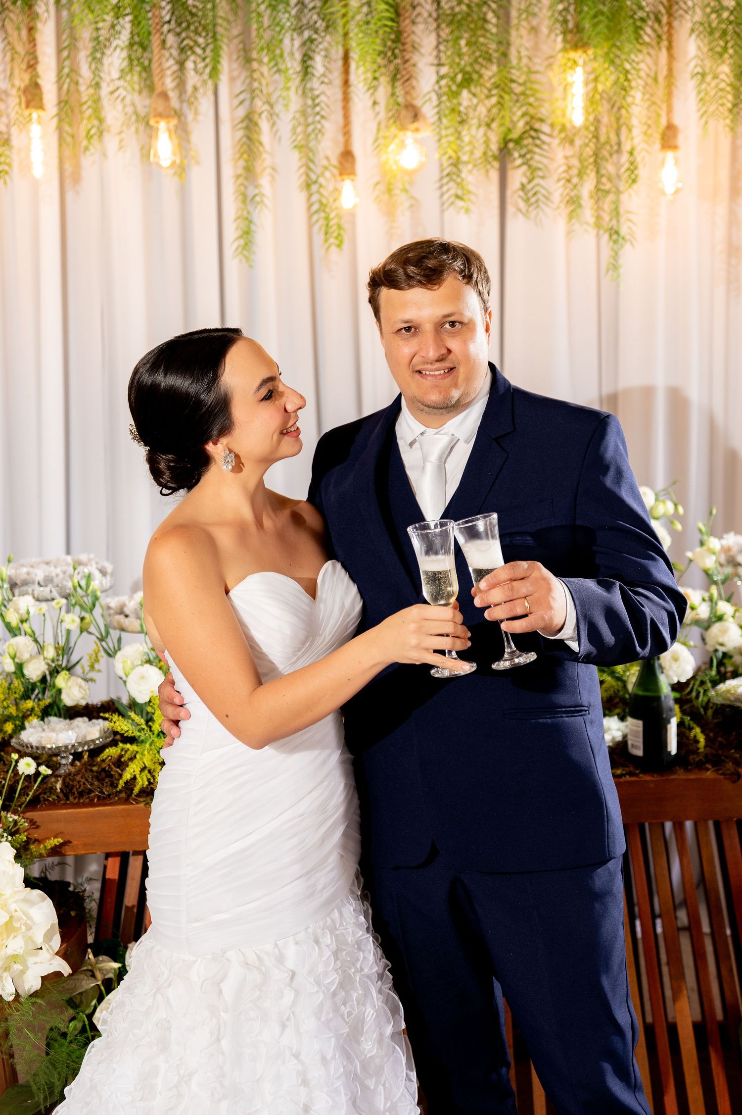 Bride and groom toasting with champagne glasses at wedding reception, decorated with greenery and warm Edison bulb lights.