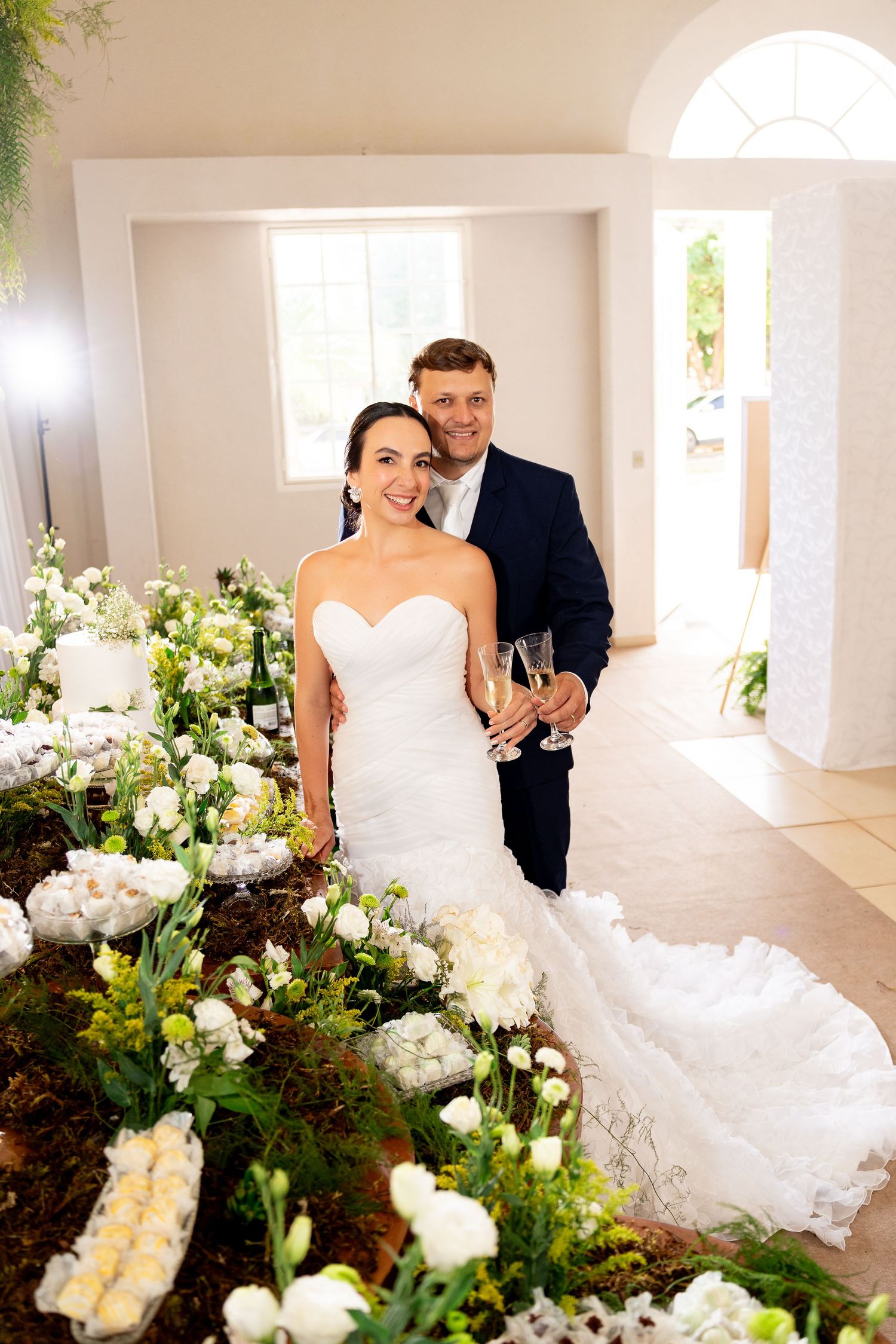Bride and groom smiling together at wedding reception, holding champagne glasses near floral arrangements and cake.