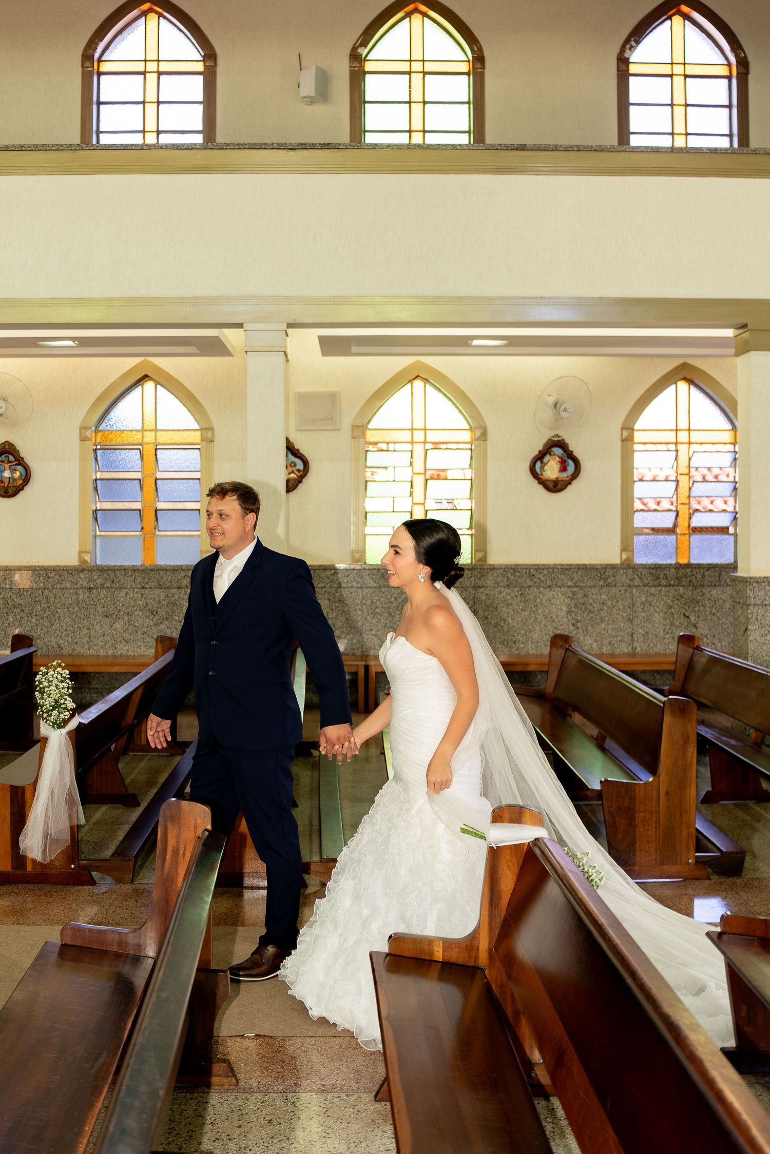 Newlywed couple holding hands walking down church aisle after wedding ceremony with stained glass windows.