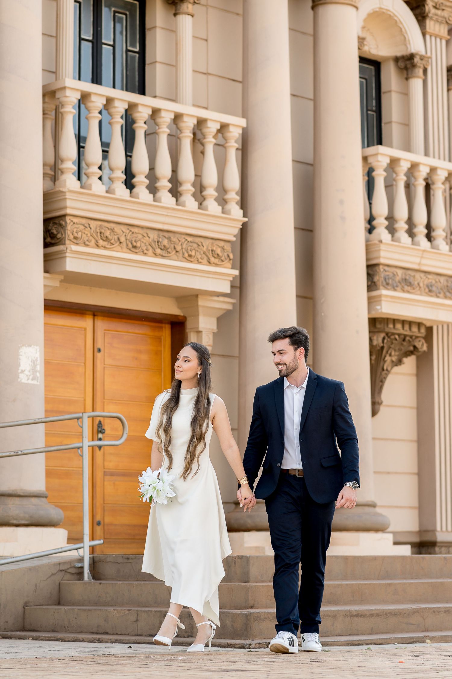 Bride and groom holding hands walking down steps of elegant classical building with ornate balustrades.